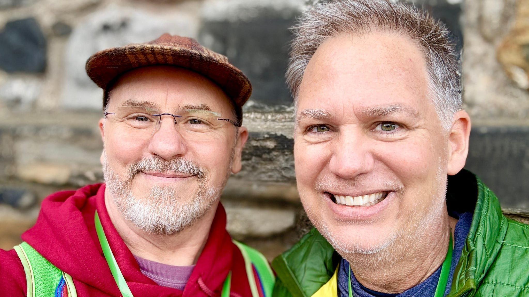 Two men are standing inside a croft house holding a sheep fleece, one of them is wearing glasses and a red hoodie and the other is dressed in a green jacket.