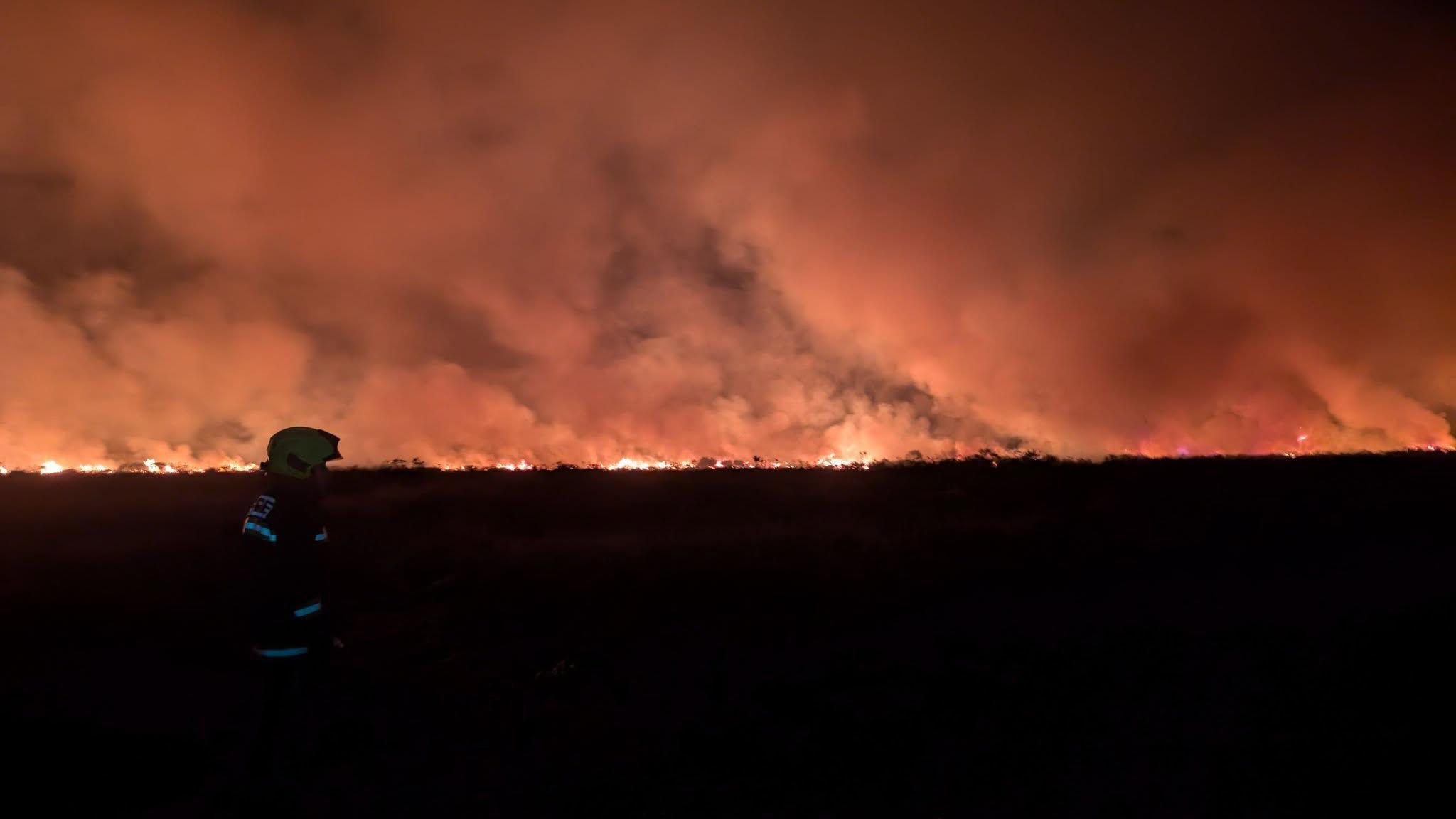 A red glow in the distance from the fires on the moorland. There is a firefighter on the left in front of the fire. It is dark but the sky is filled with smoke. 