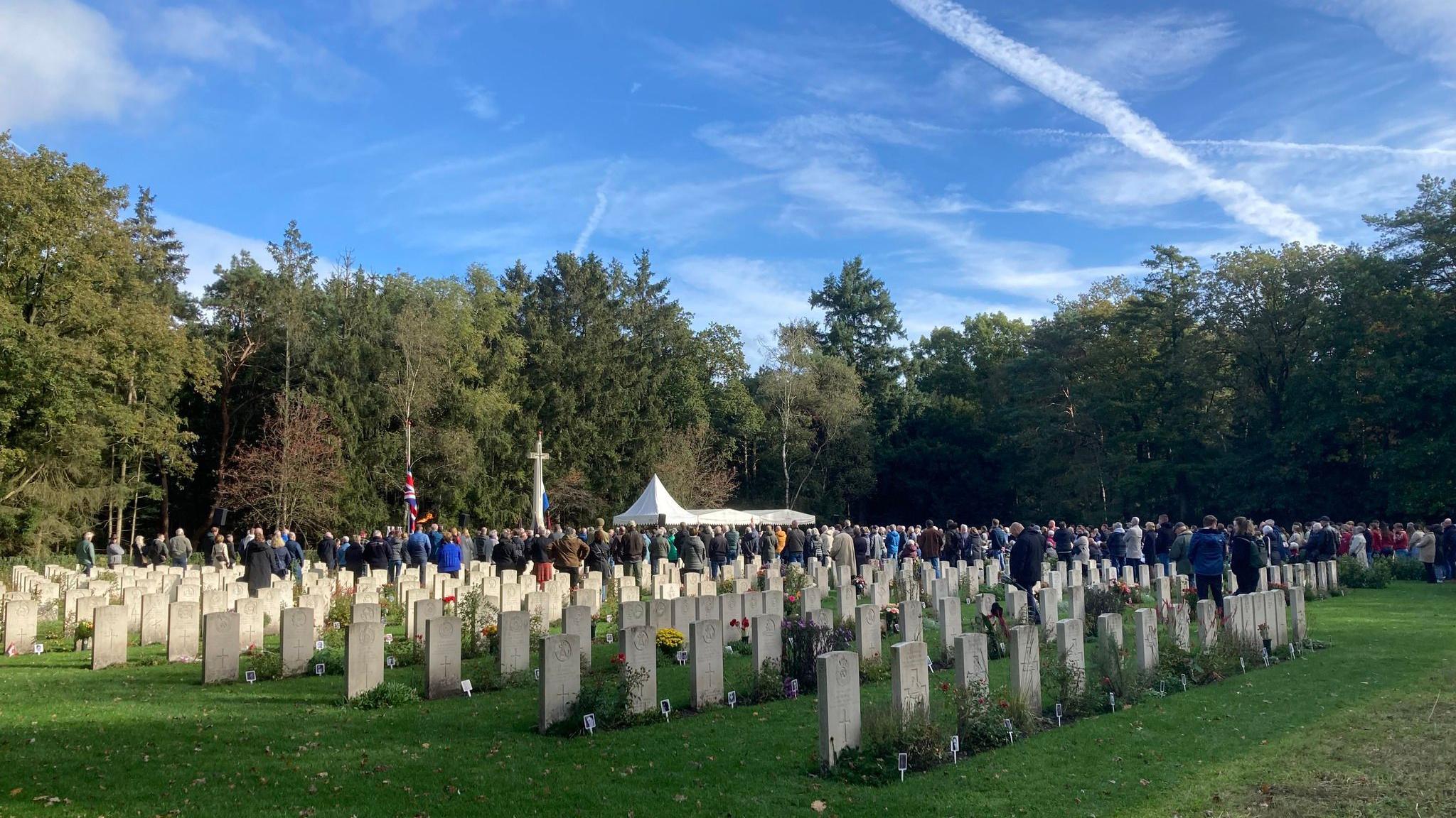 Grey stone graves in a war cemetery with flowers - in the distance people stand around a white marquee with flags and a large cross