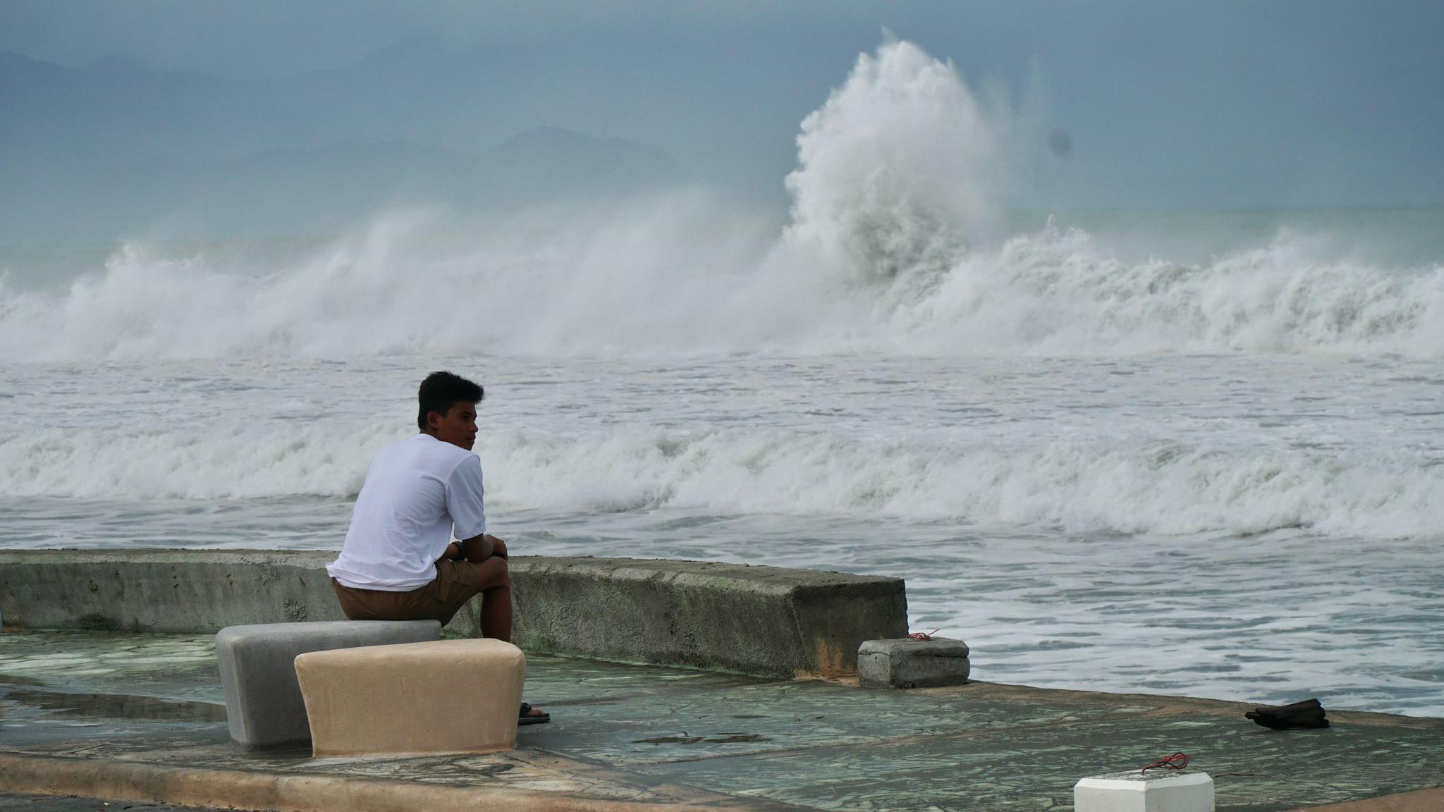 A man sits looking out to sea on a concrete bench as large foaming waves roll in.