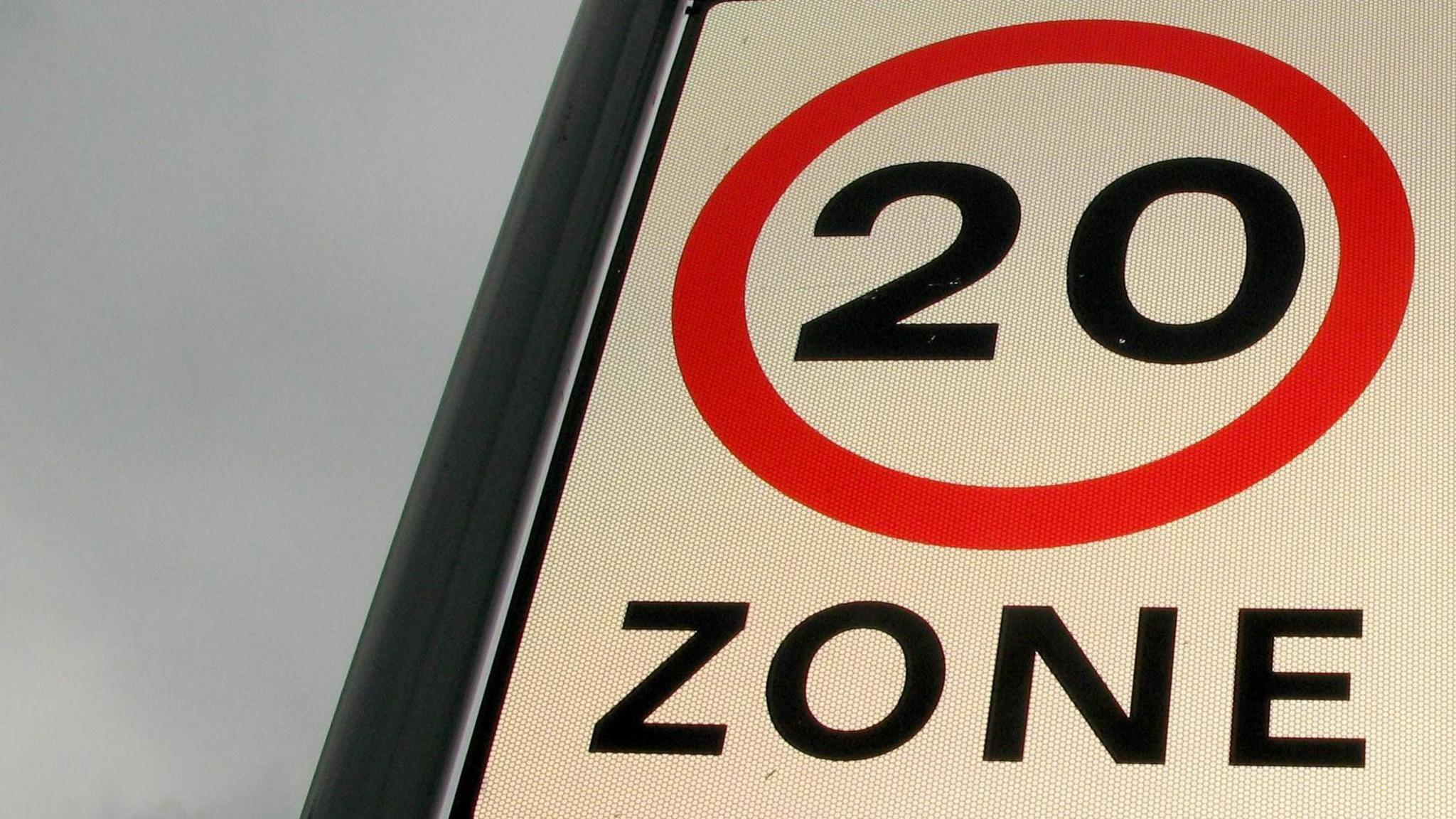 A road sign with the number 20 and the word "zone" in black writing on a white background. There is a red circle around the number, and a cloudy sky in the background.