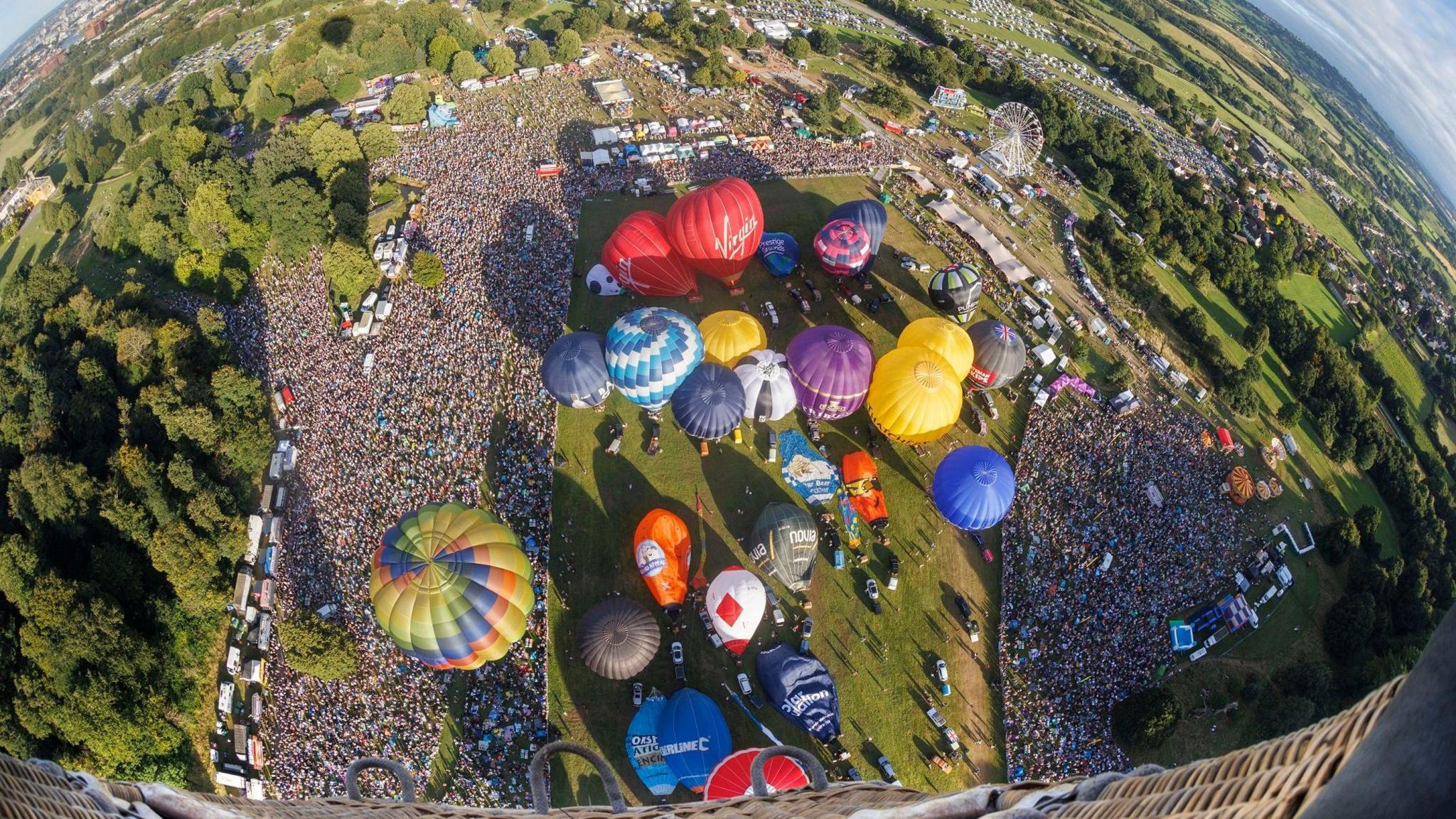 The site of the Bristol Balloon Fiesta is seen from the basket of a hot air balloon that has just taken off. Thousands of spectators can be seen on the ground, along with dozens of other balloons which are inflated ready to take off. Fields and the boundary of the city of Bristol can also be seen in the distance