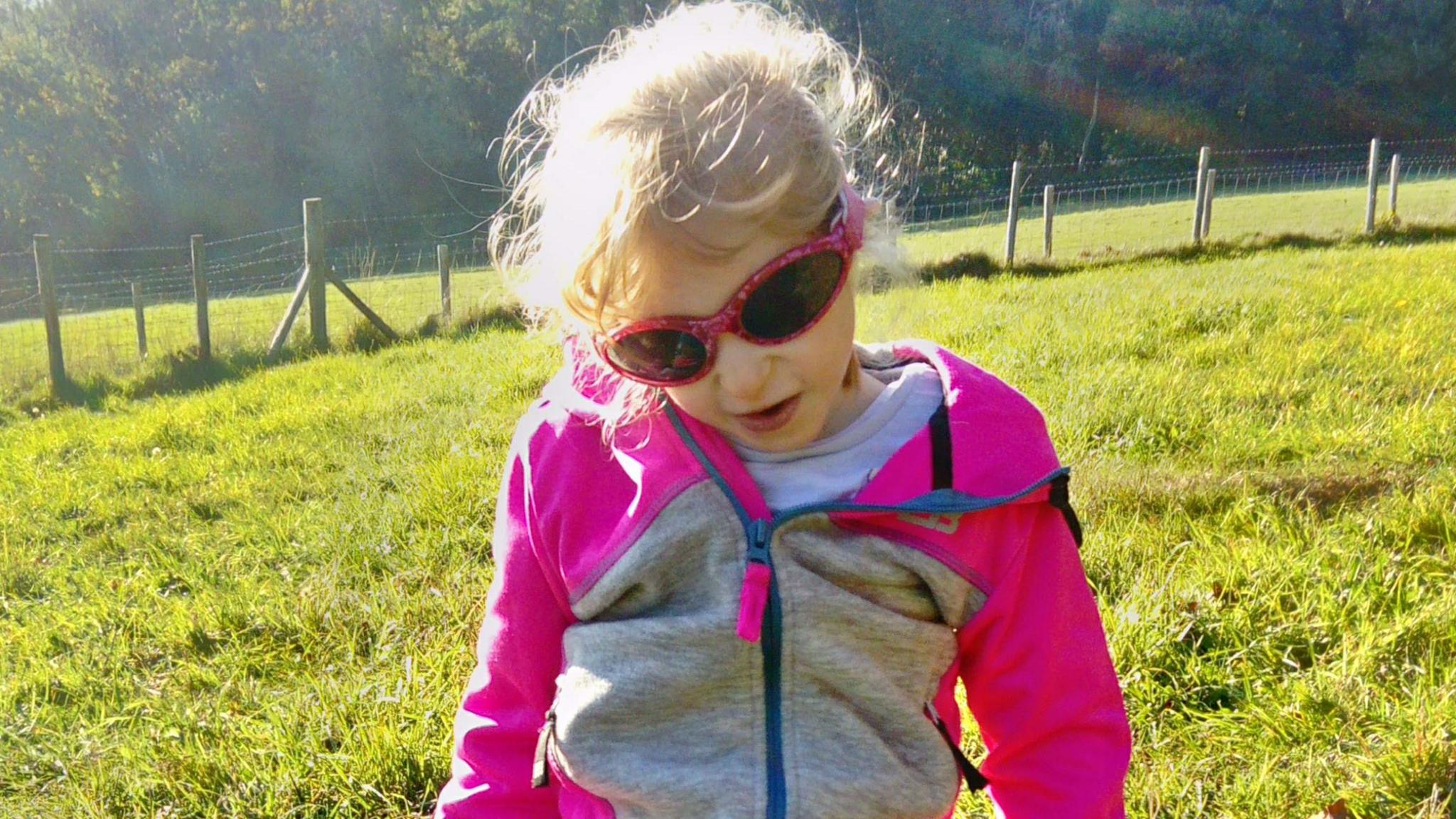 Jaydi wearing a pink and grey zip up hoodie with pink sunglasses while out on a sunny day in a grassy field with trees in the background