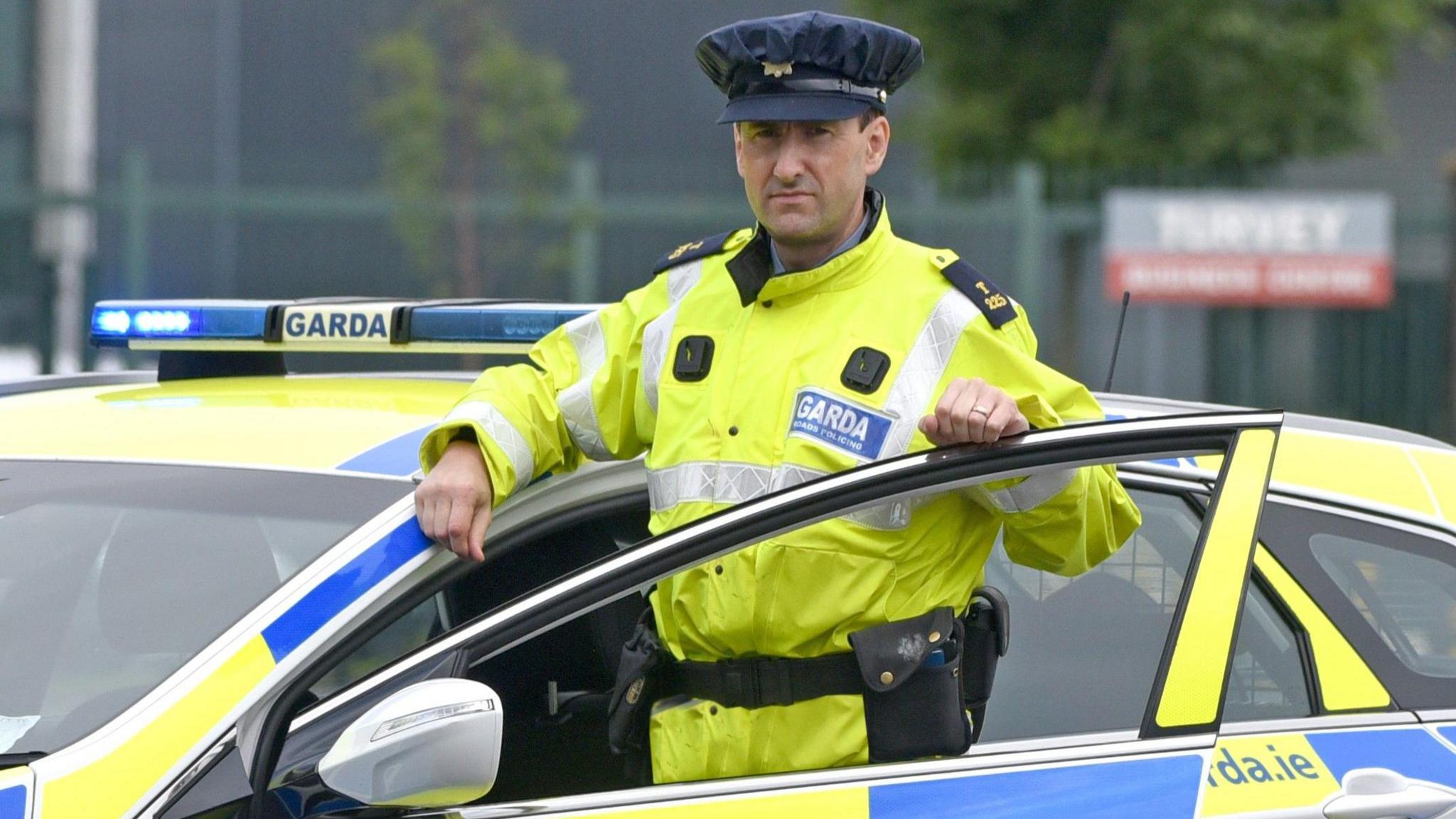 A man stands in-between a police car door and its body. He wears a navy hat and yellow illuminous vest which has the words 'Garda' on it in silver font with a blue background.