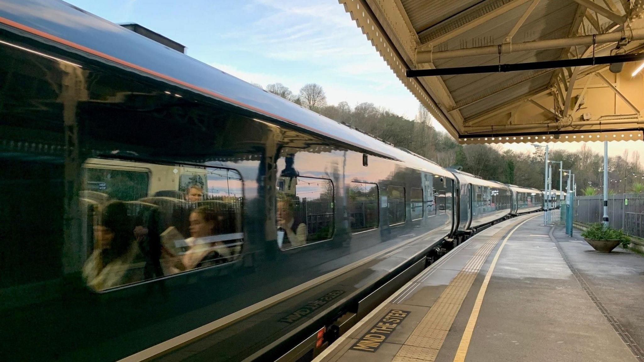 A dark Green Great Western Railway train departs Bath Spa station. Passengers can be seen sitting inside the train.