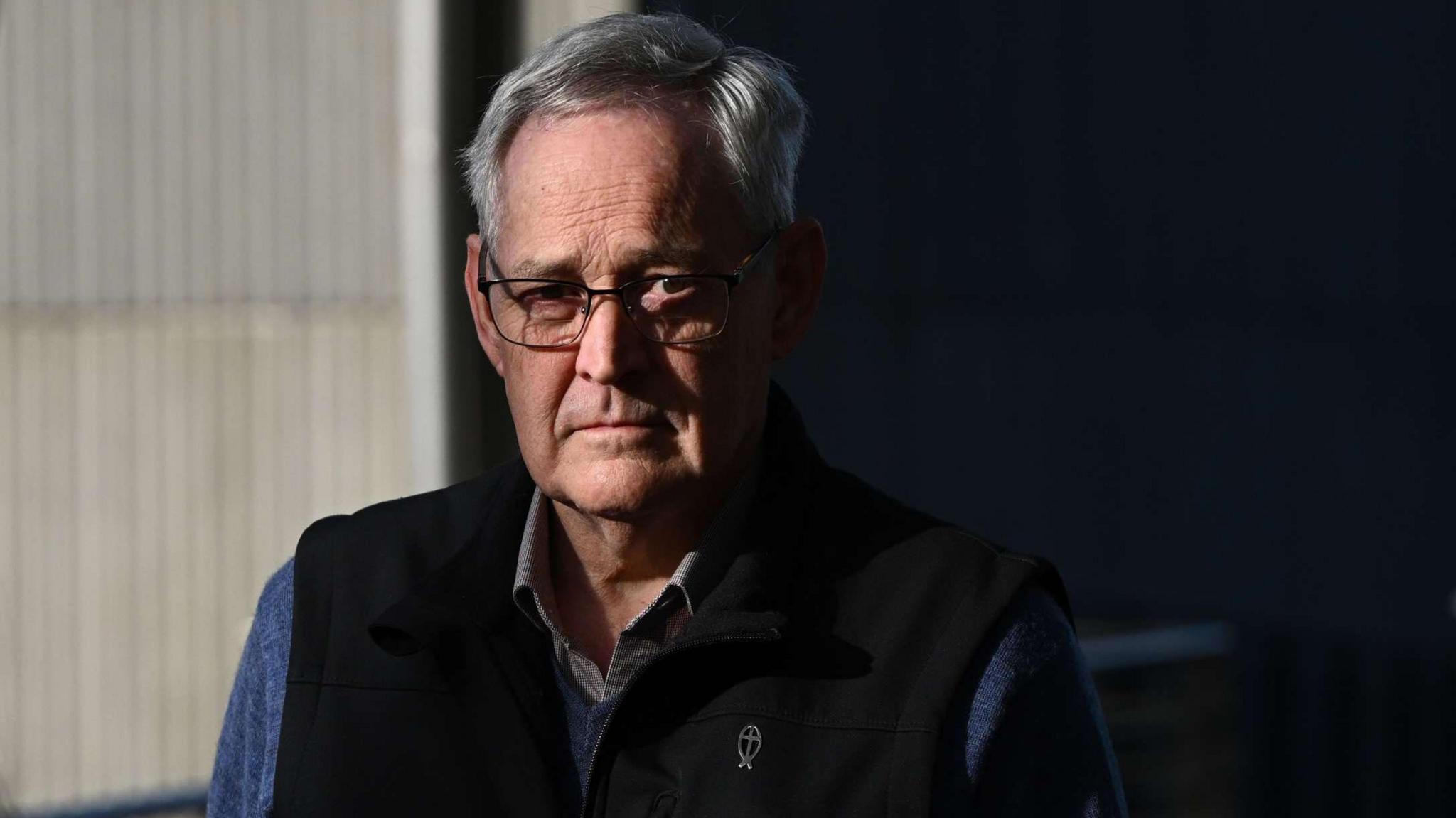 Ian Wilkinson with white hair and glasses looks at the camera. He wears a blue shirt and a  black vest.