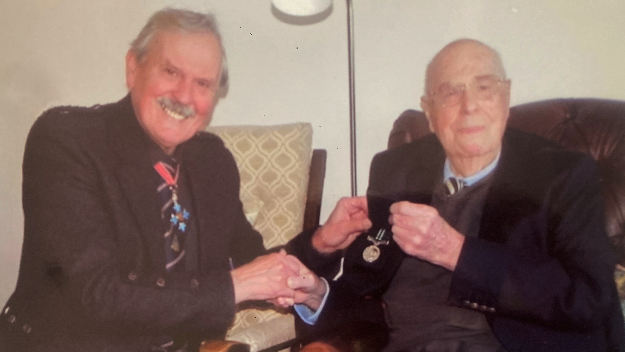 John Cruickshank and Bob Kemp are sitting in armchairs next to each other and are smiling at the camera. Both are wearing ties and blazers. John has a bald head and glasses and Bob has grey hair and a grey moustache. Bob is wearing a medal and is presenting John with a medal.