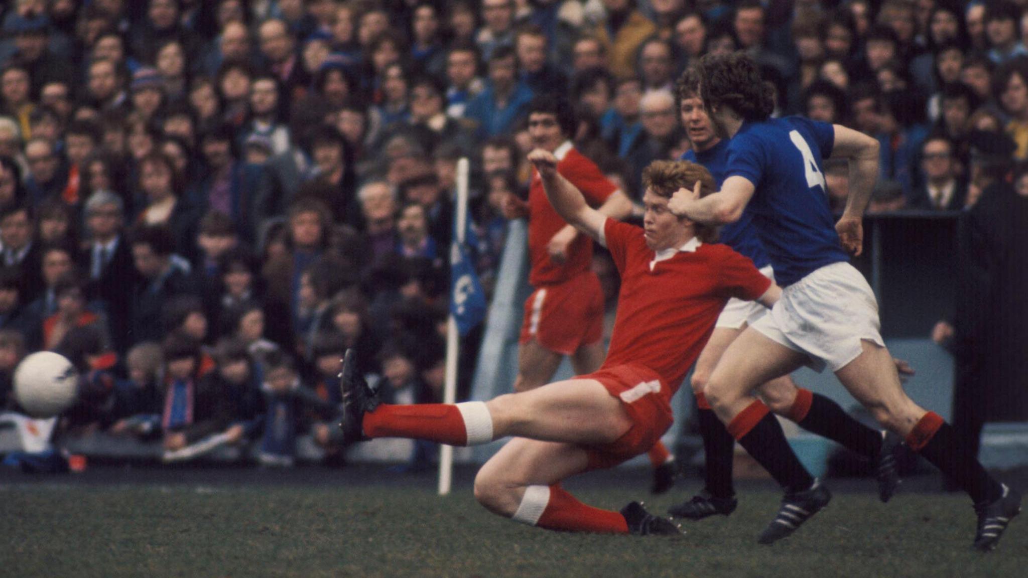 Willie Young wearing a red Aberdeen shirt tackling a Rangers player wearing a blue shirt with white shorts and black socks.