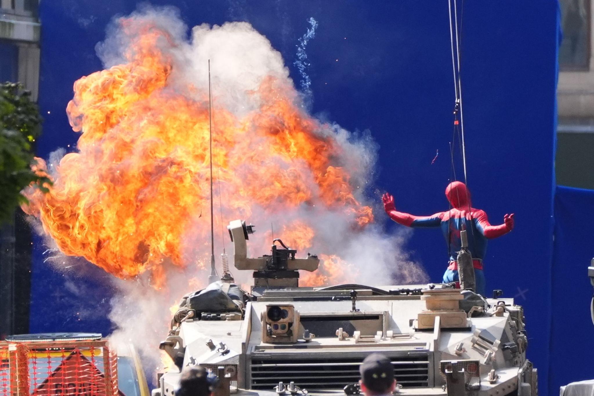 Spider-man on a tank gestures towards a controlled explosion during filming. 