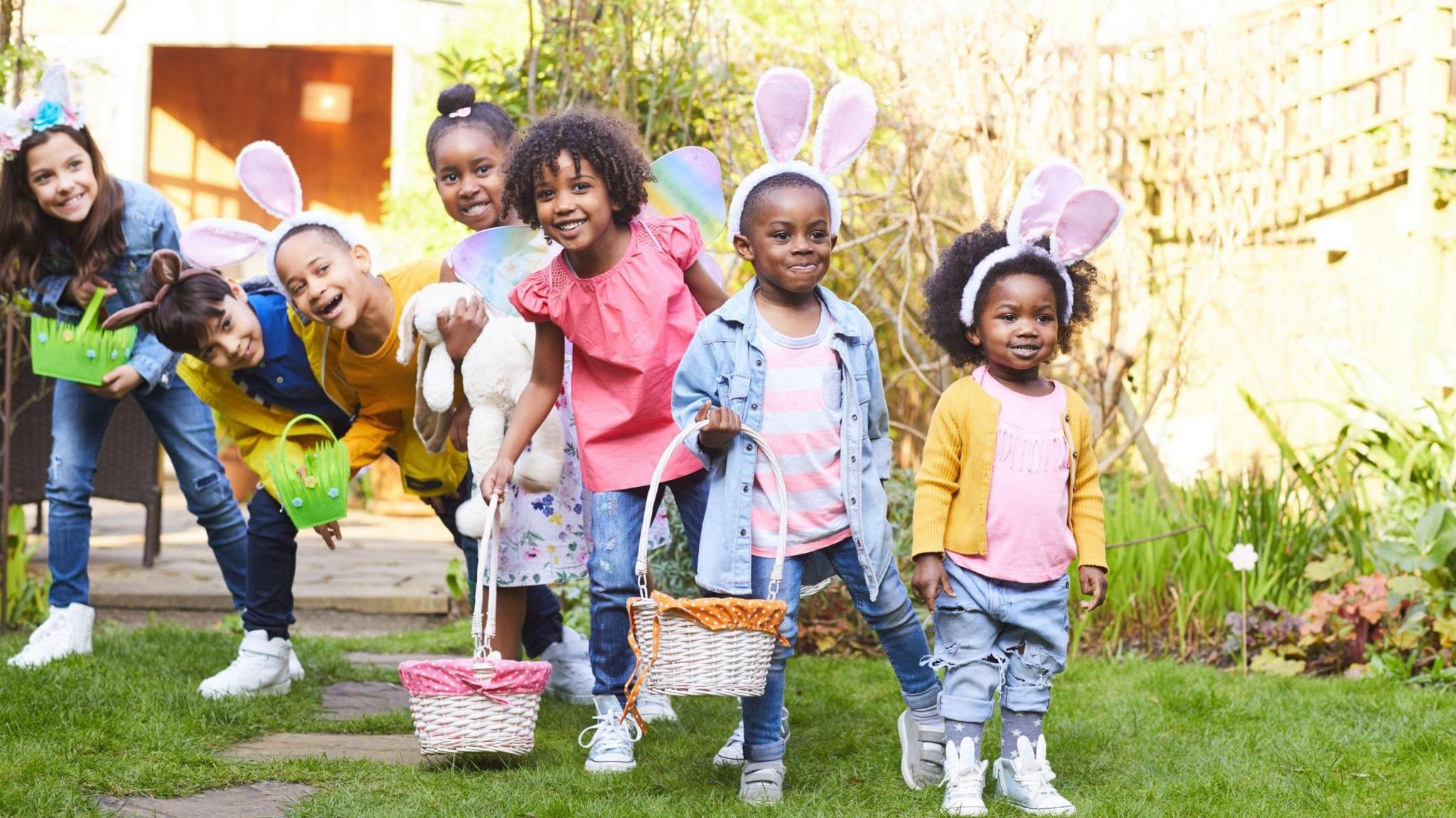 Seven children lined up in a row in a garden wearing bunny ears and holding easter egg baskets. They stand on grass in front of a fence with plants in front of it.