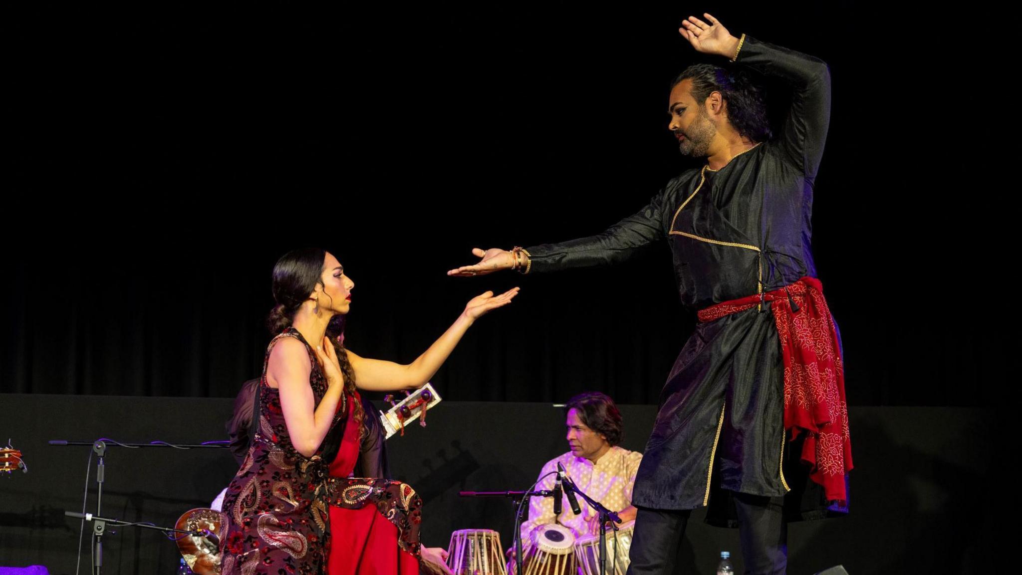 Two dancers on the stage, both are performing Kathak while wearing traditional Indian clothes. A tabla (drums) player is in the background 