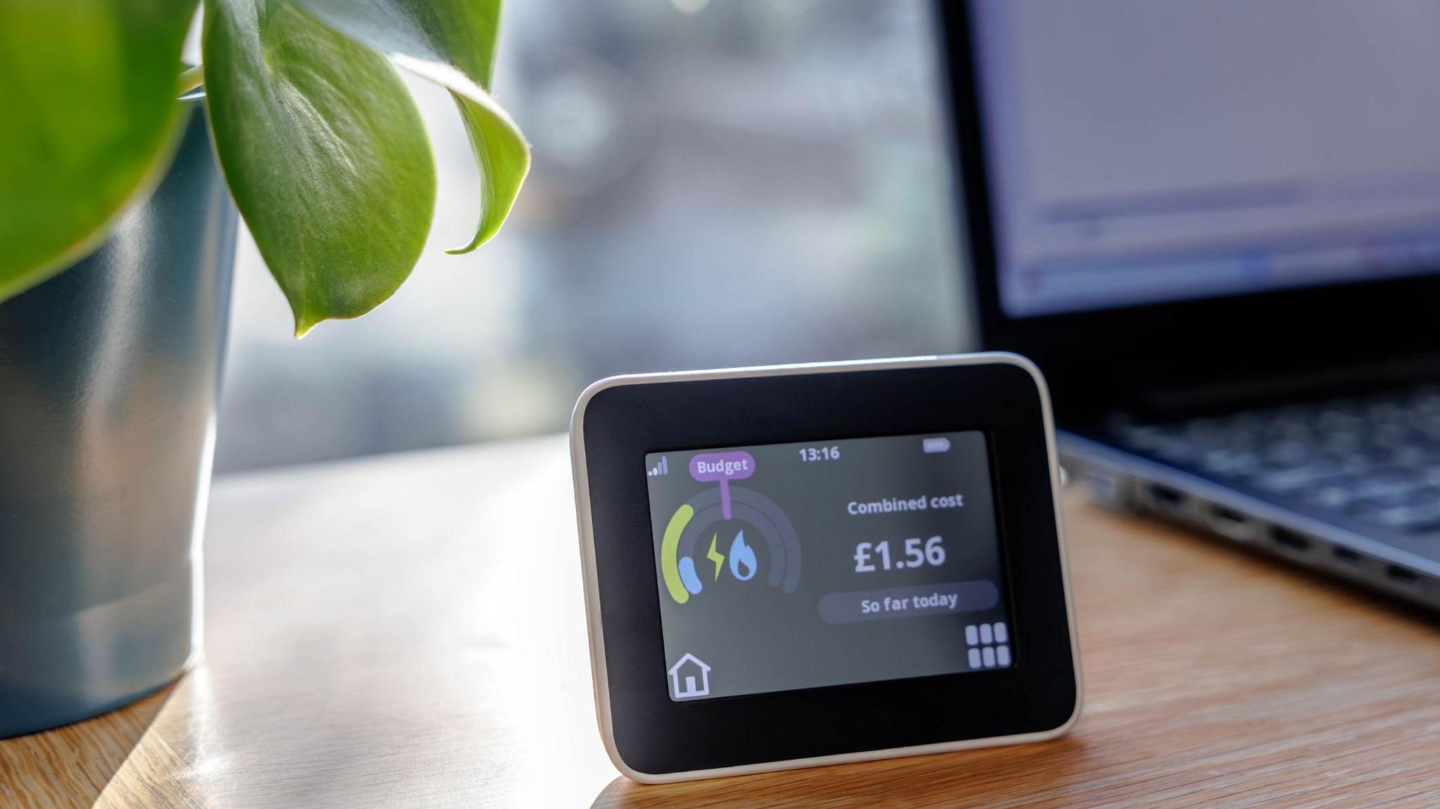 a domestic energy smart meter showing a daily cost of £1.56 is sitting on a table with a laptop computer and a potted plant beside it