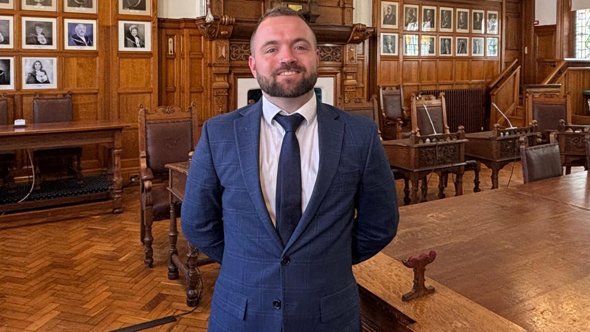 Councillor Devon Watson, who has short brown hair and a beard and is wearing a navy suit. He is smiling proudly as he stands with his hands behind his back in the Douglas Council chamber. 