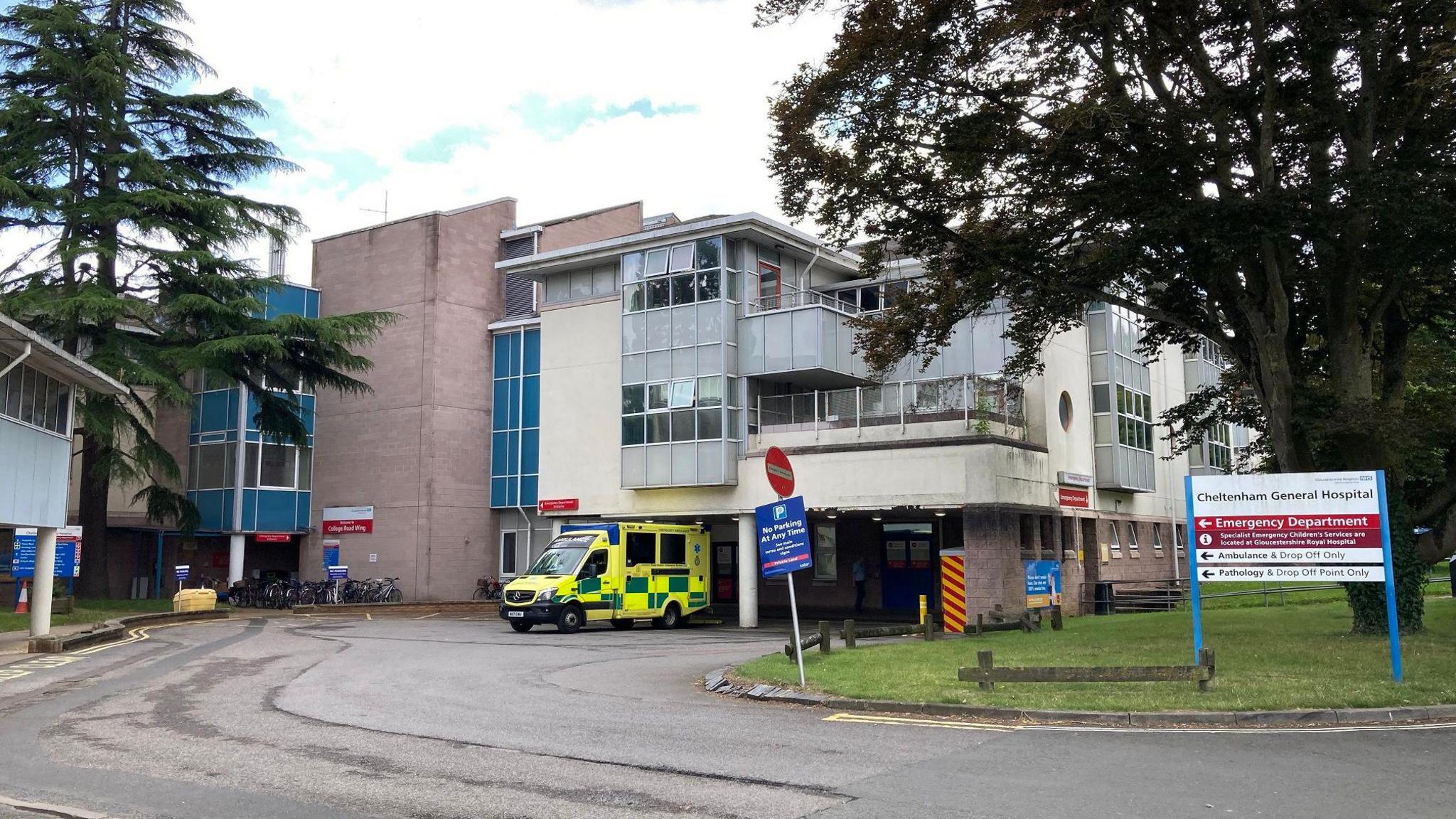 The exterior of Cheltenham General Hospital in Gloucestershire. It is a largely grey building with some cream panels, and there is one ambulance parked outside