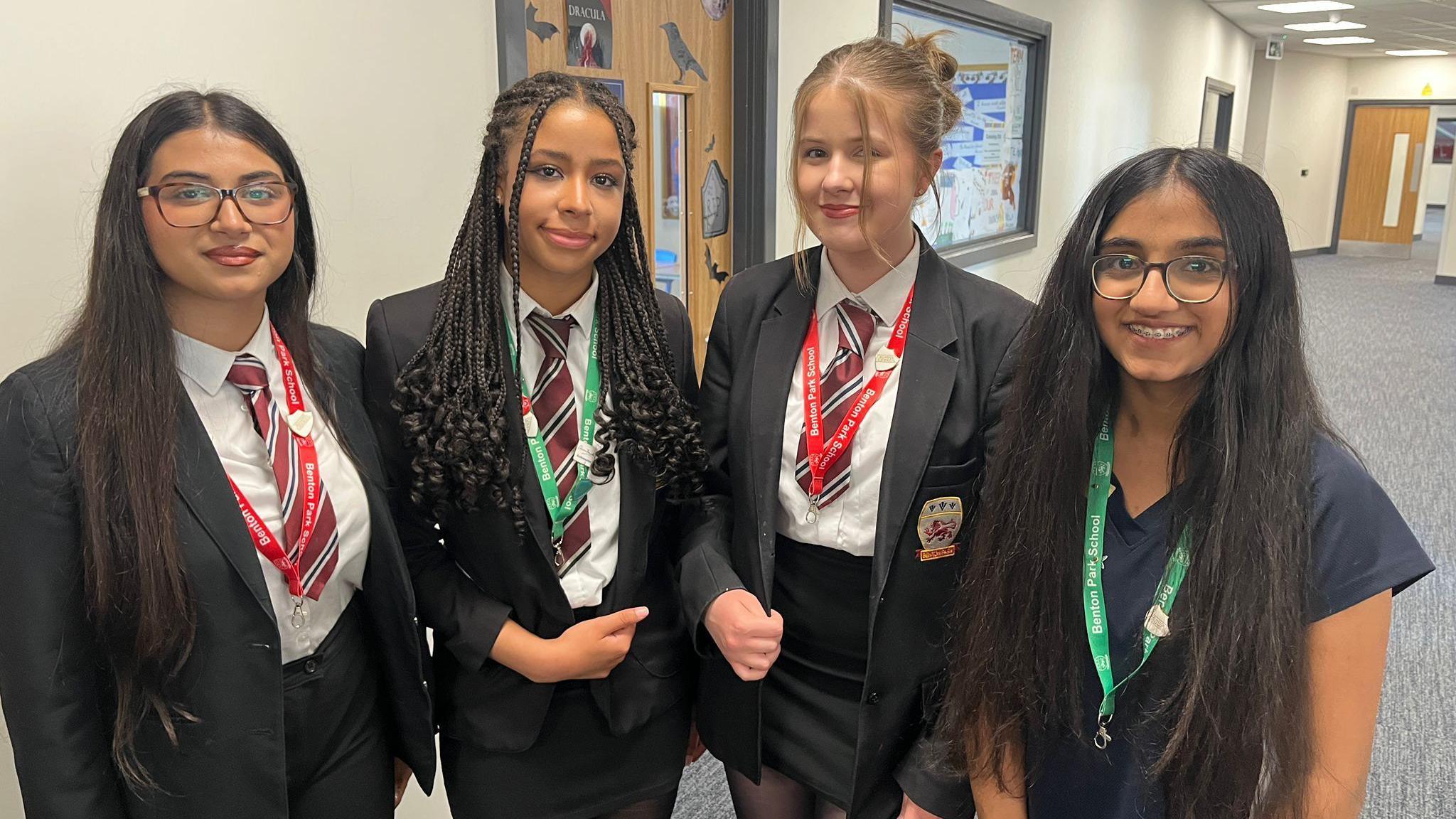 Four teenagers stand beside each other in a school hall. Three are wearing black blazers, black school skirts and red, navy and white stripped ties and are smiling. The fourth girl is wearing a navy t-shirt and glasses.