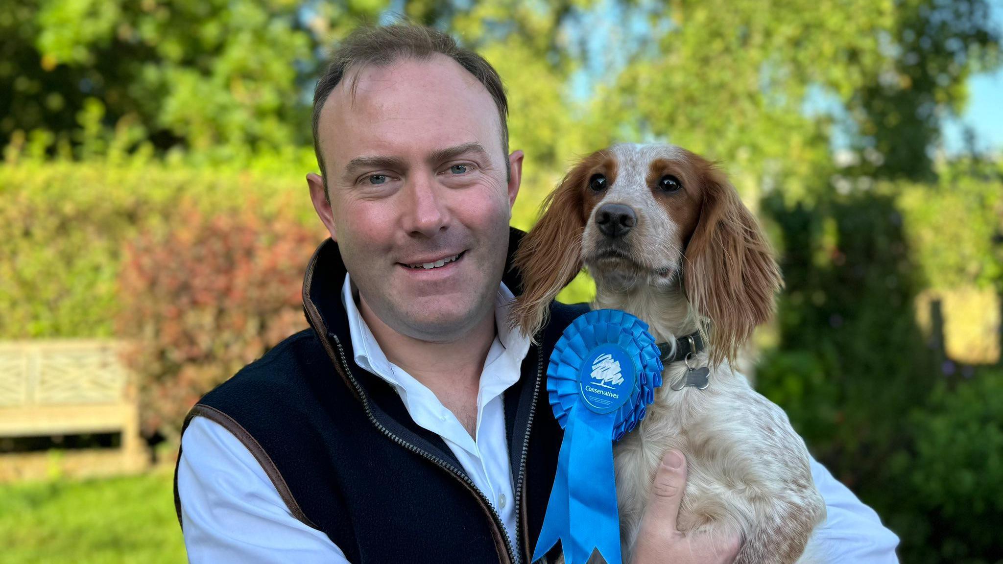 A smiling Blake Stephenson looking directly into the camera, wearing a gilet over a white shirt and holding a dog who has a Conservative rosette attached to its collar. 