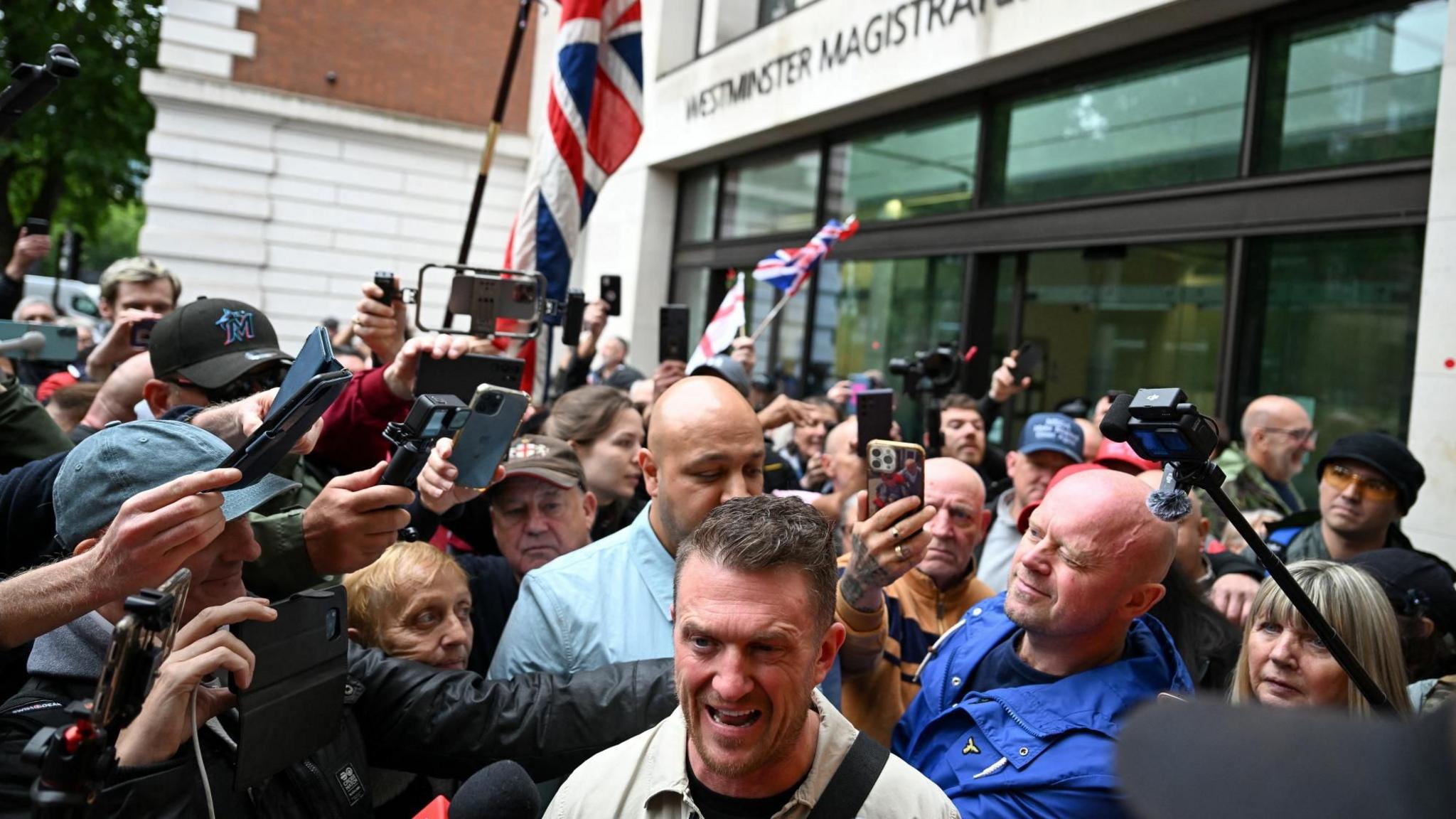 Tommy Robinson/Stephen Yaxley-Lennon in the foreground with a scrum of reporters holding up phones and microphones on his right. His supporter are behind him holding a couple of Union or England flags. The glass entrance doors of Westminster Magistrates' Court are behind them all.