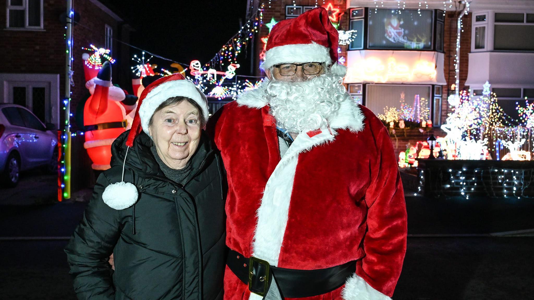 A woman wearing a black coat and a Santa hat stands next to a man wearing glasses and a full Santa outfit - including beard. Behind them is a house covered in bright lights.