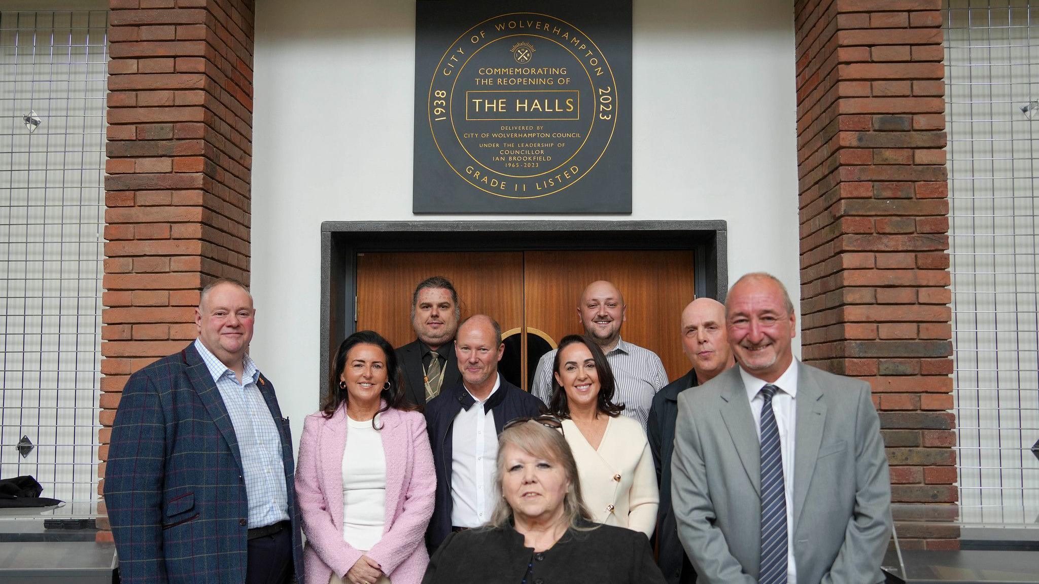 Nine people assemble for a photo in front of a door, above which is a black-coloured square plaque with golden writing. 