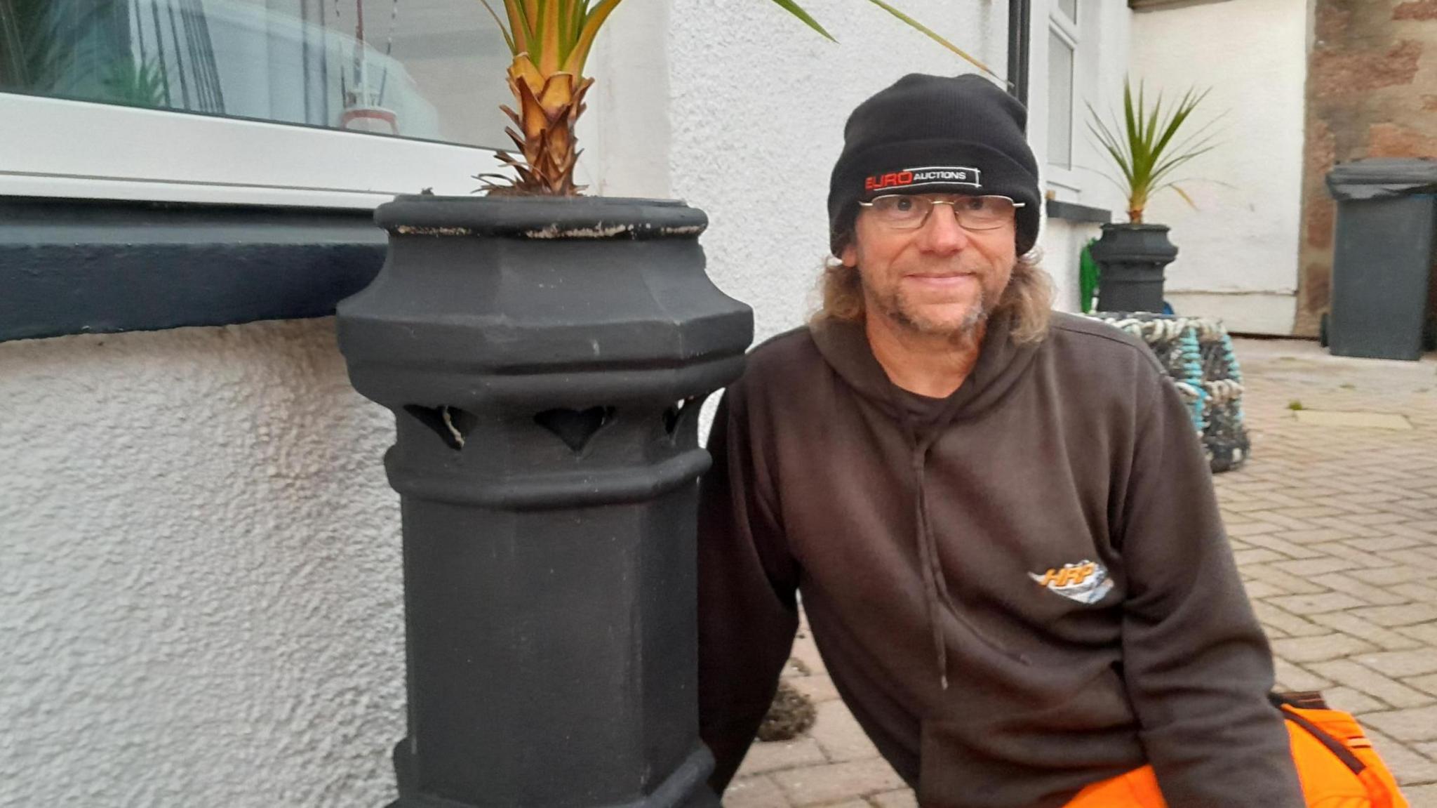 Ian Suddaby is wearing a black hat and glasses and smiling. He is wearing a dark hooded top and orange trousers. He is sitting beside a chimney pot.