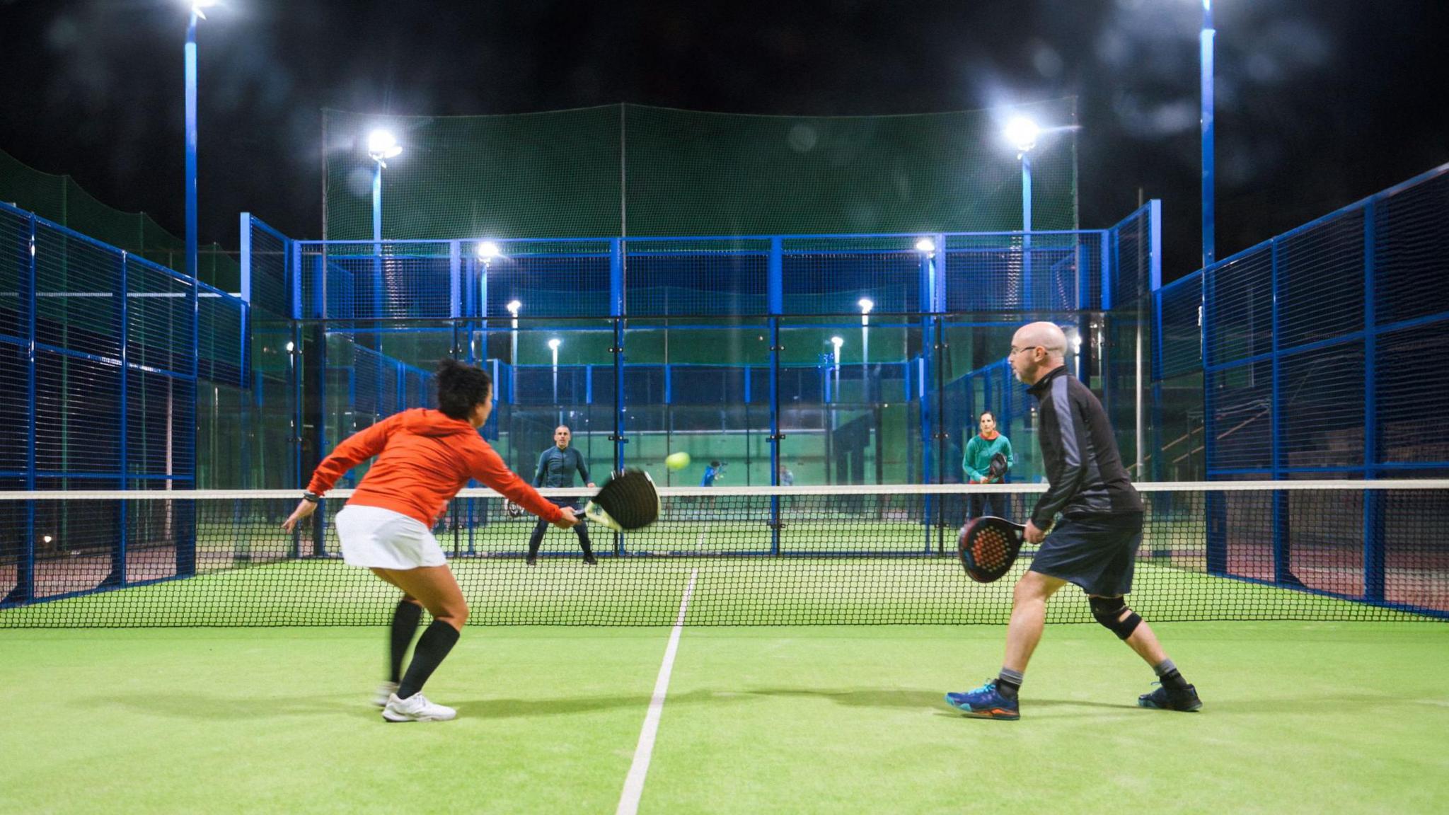 Four people playing the raquet sport padel at night on an artificial grass court. There are two people on one side of the net, one wearing an orange hoodie with white shorts and another with a black jacket and blue shorts. There are two others facing them.