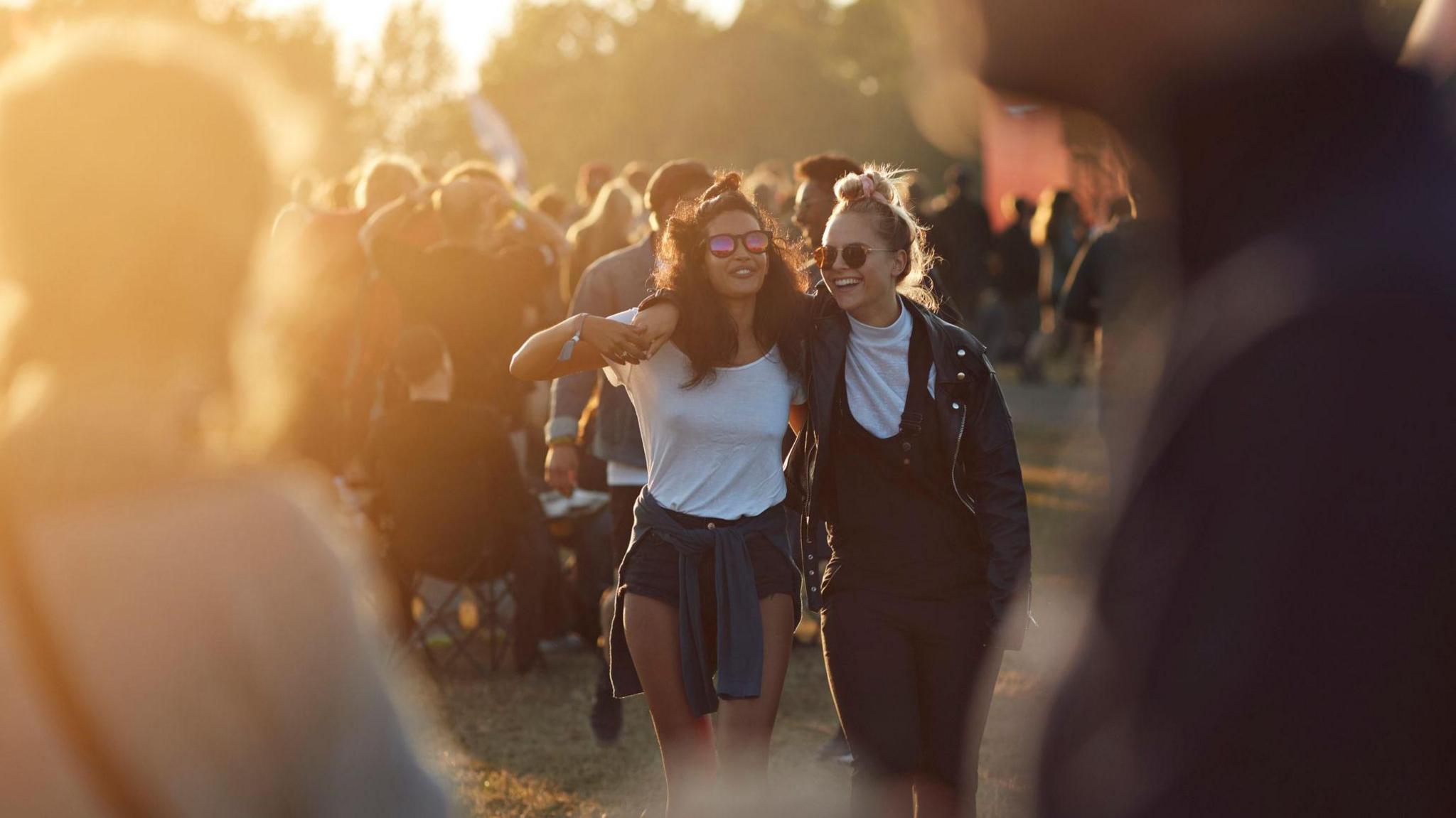 Two girls walking through a festival at dusk, with their arms around each others shoulders and both wearing sunglasses.