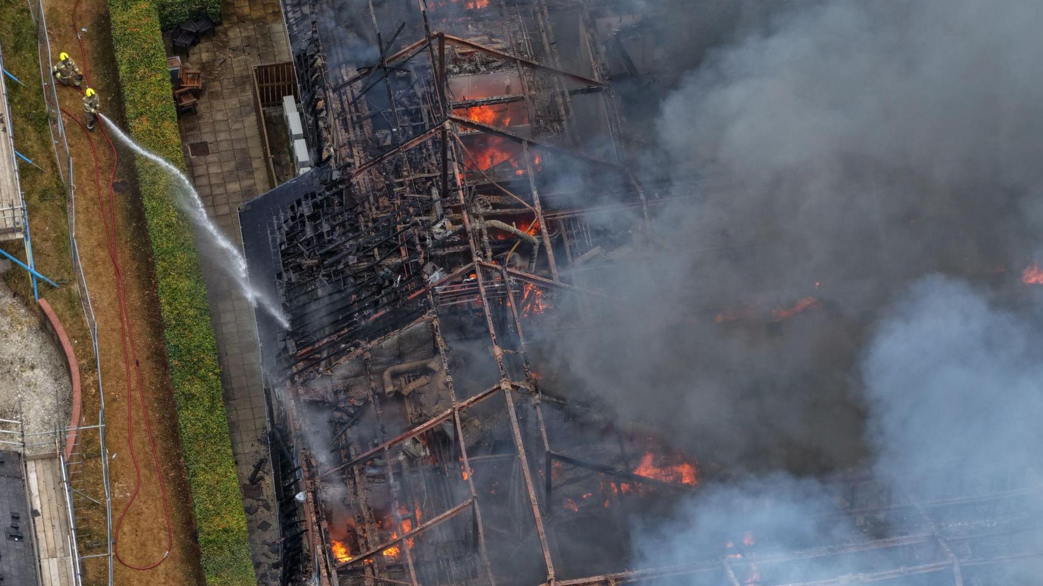 An aerial view of a large building engulfed in flames, with its roof collapsed and thick smoke rising. Two firefighters on the ground spray water from a hose toward the burning structure, surrounded by a paved walkway and hedges.