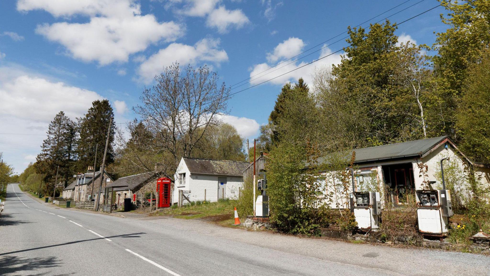 The main street at Calvine. A small garage and three stone houses run along the side of the empty road. There is an old-fashioned red phone box and a similarly traditional post box can be seen in the wall of one of the buildings.