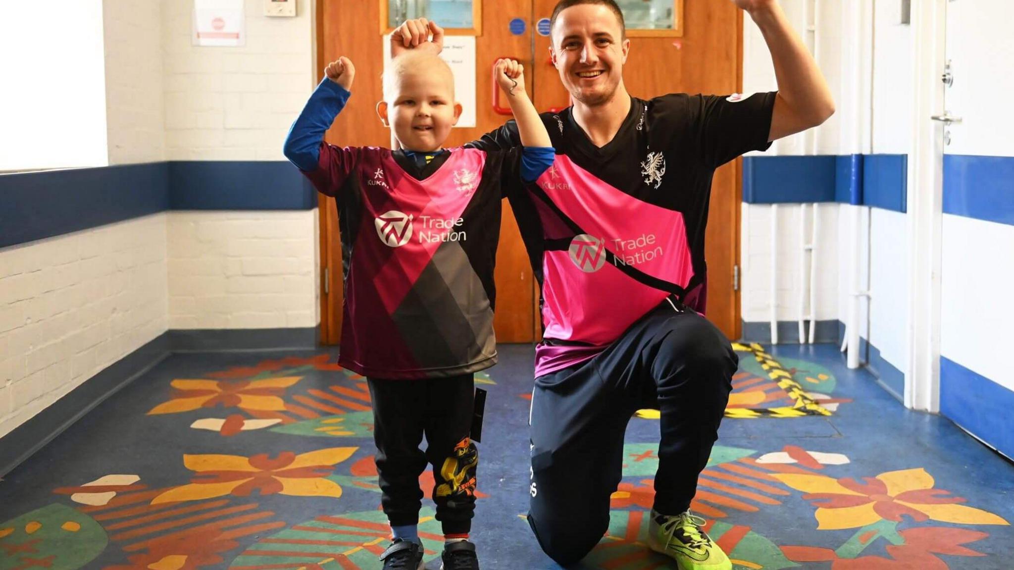 Bodhi is a young boy and is standing in a corridor next to a cricket player wearing the team's pink and black shirt. Both have their fists raised in the air, smiling at the camera.