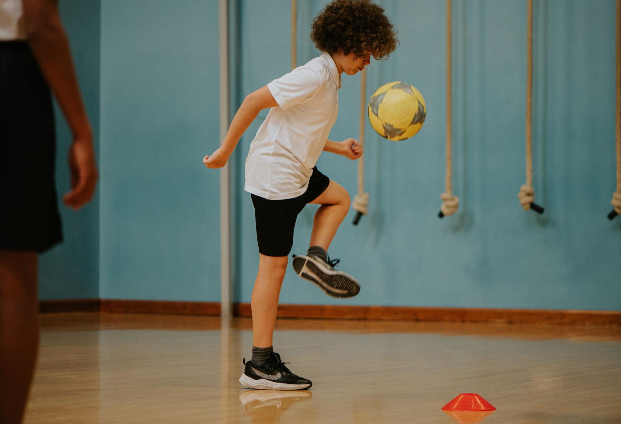 A boy with curly brown hair does a keepy-uppy in a school gym