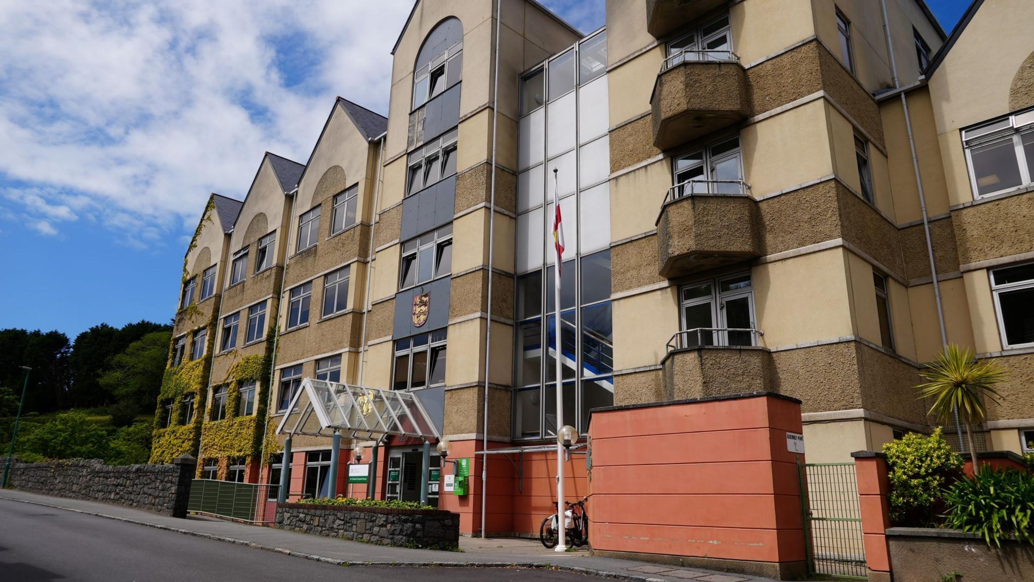 A four-storey government building with concrete walls painted yellow, pebble dash stripes, ivy growing up some parts and gabled roofs.