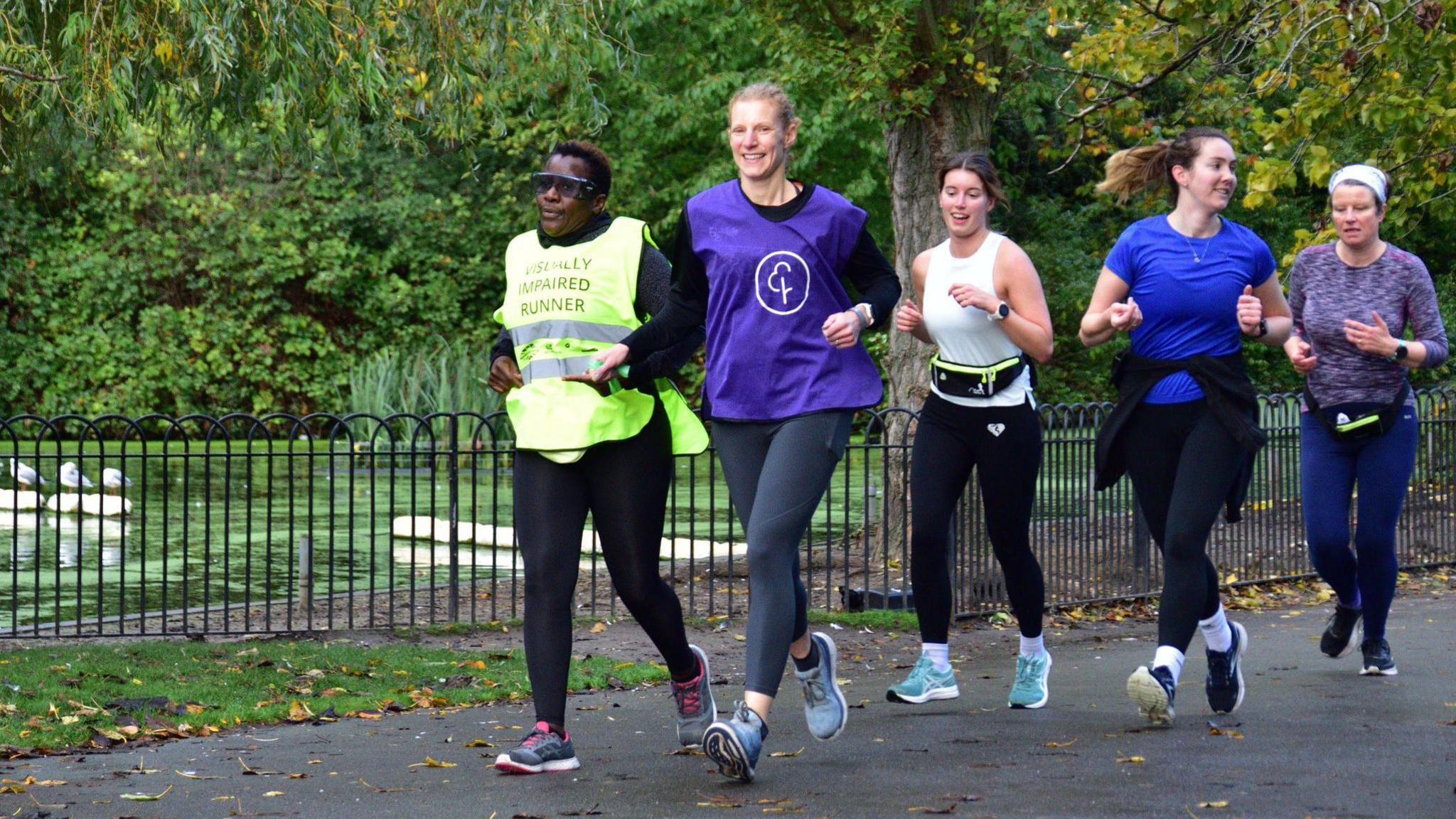 Judith Namaganda, wearing a visually impaired runner bib, running by a pond in a park with four other female runners, one of whom is her guide