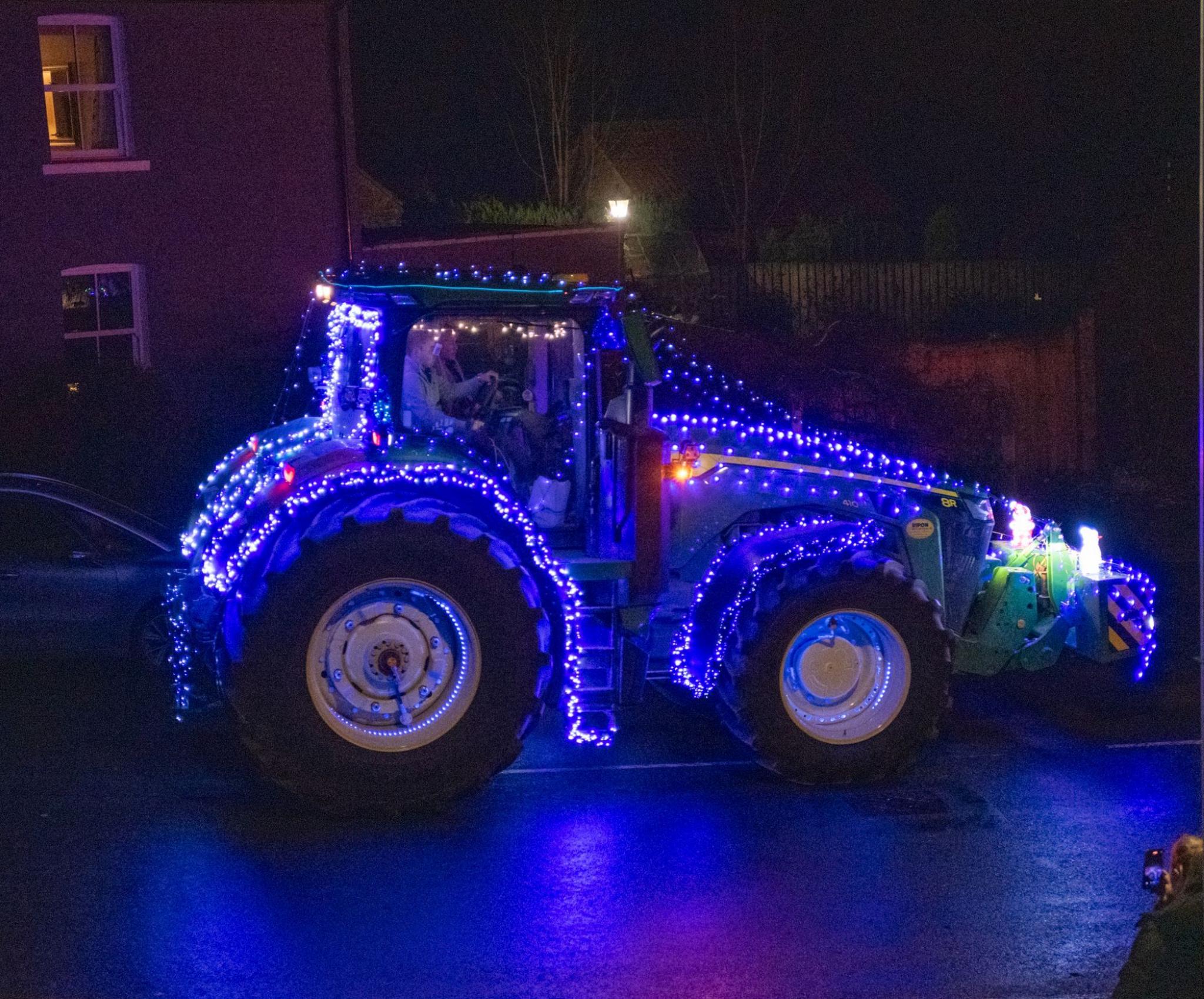 A tractor covered in blue string lights makes its way through a village.