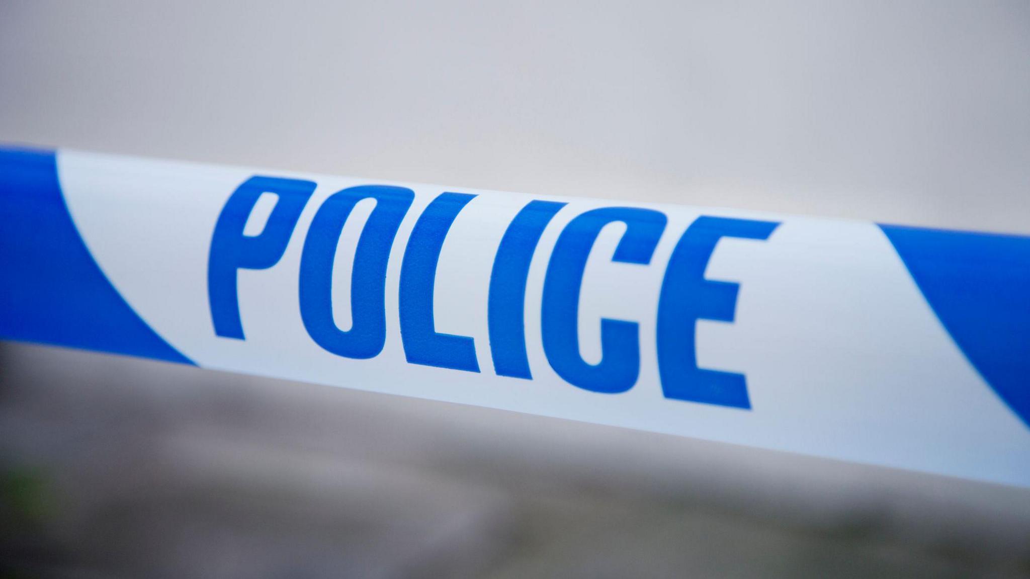 A stock image of some police tape with blue writing on a white background. The background is blurred with no identifiable features.
