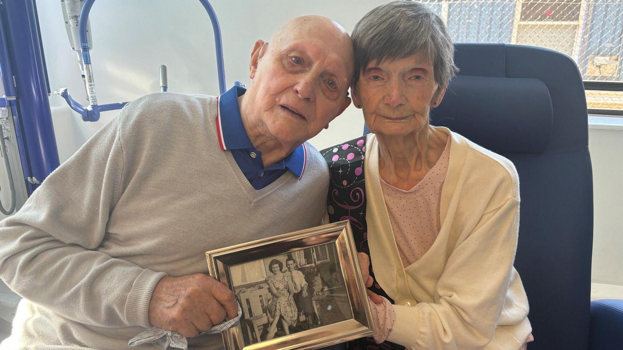 Elderly woman sits on hospital chair next to elderly man. The couple are holding black and white photo of themselves as a much younger couple. 