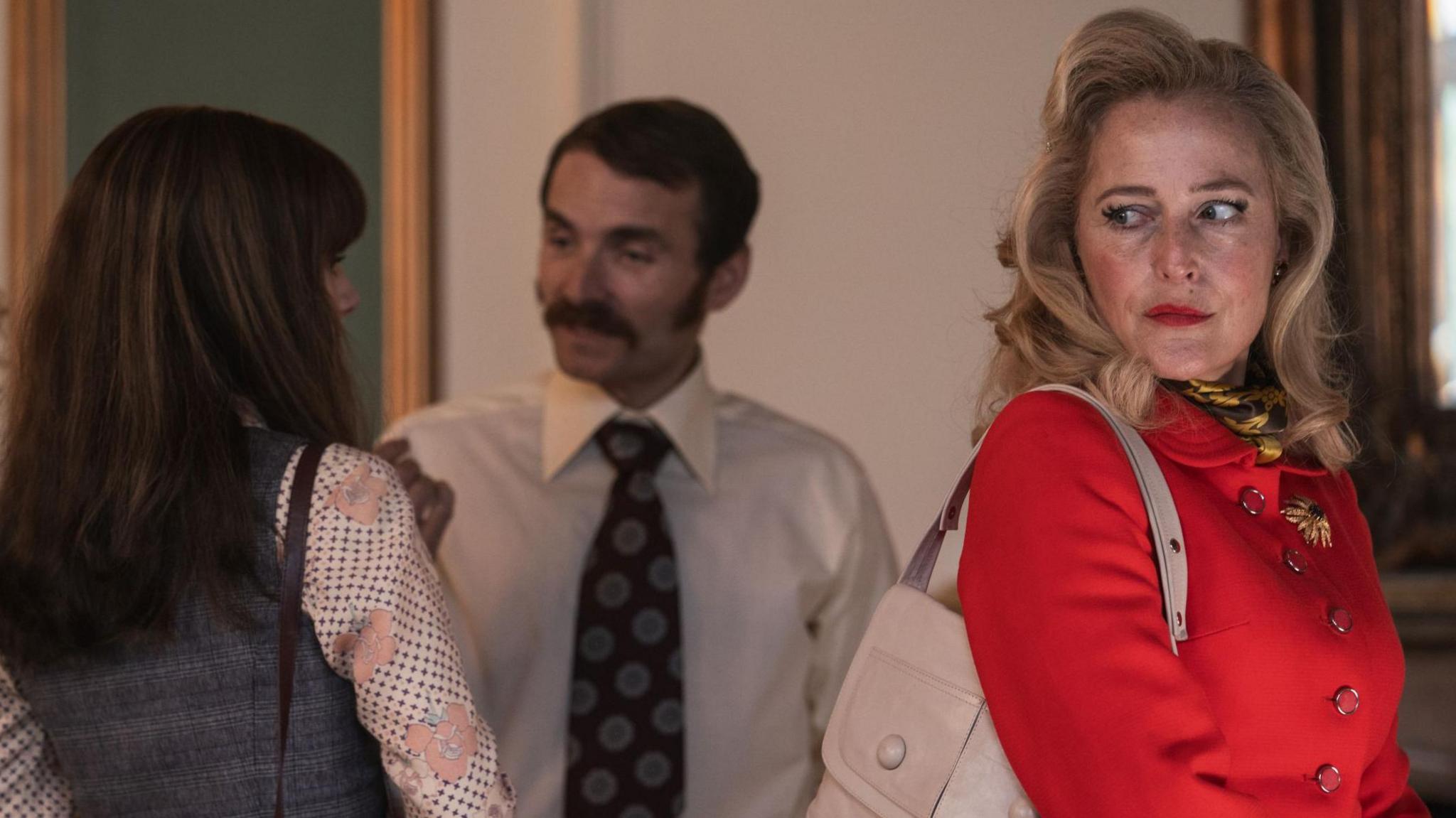 A still from the TV show Trespasses. A woman with long blonde hair wearing a red buttoned up blazer is on the right. She is looking over her shoulder with an annoyed expression. Behind her a man in a shirt and tie is talking to a young woman with long dark hair.