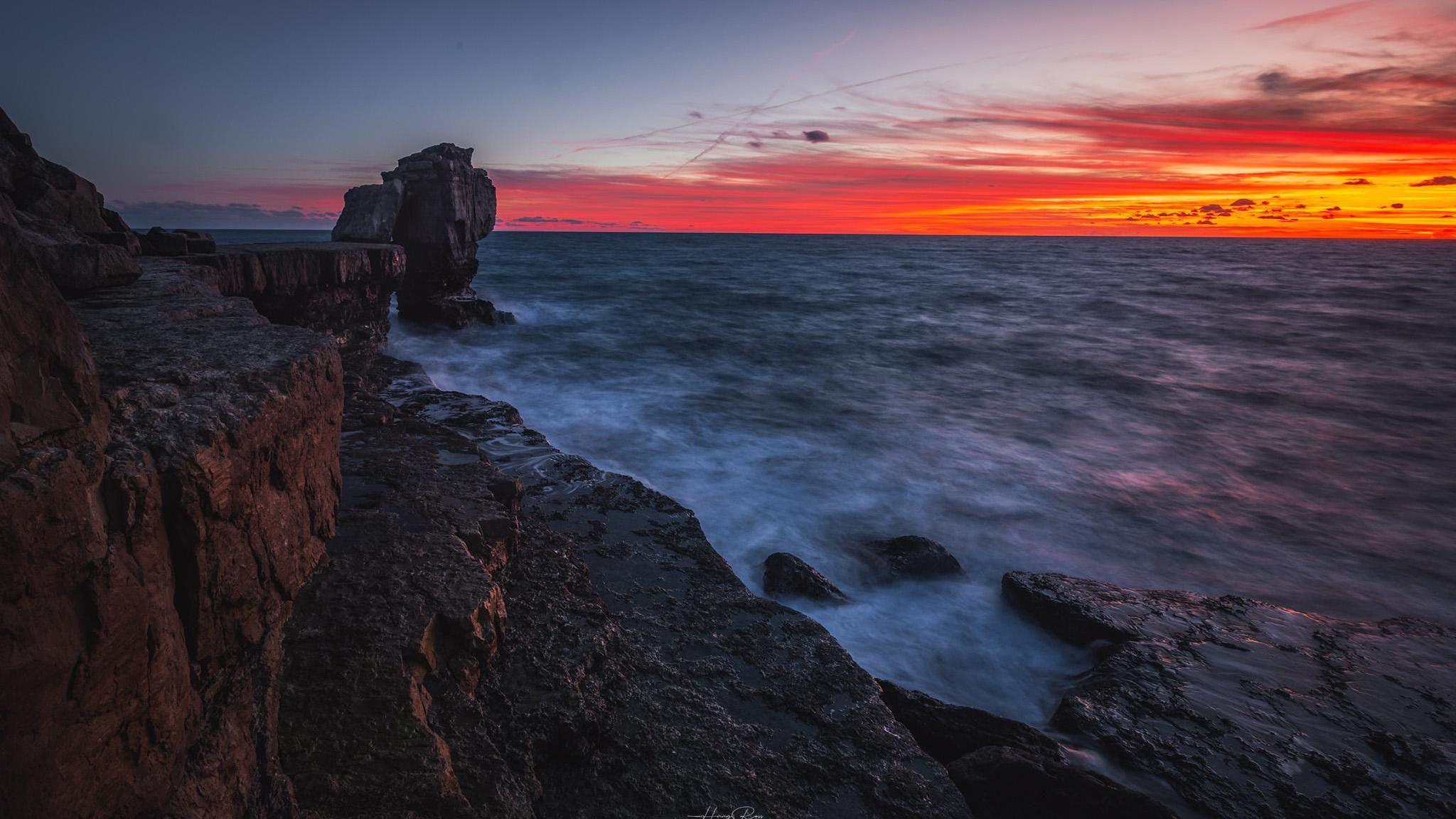 A bright red sunset overlooks a choppy looking sea in front of a rocky coastline