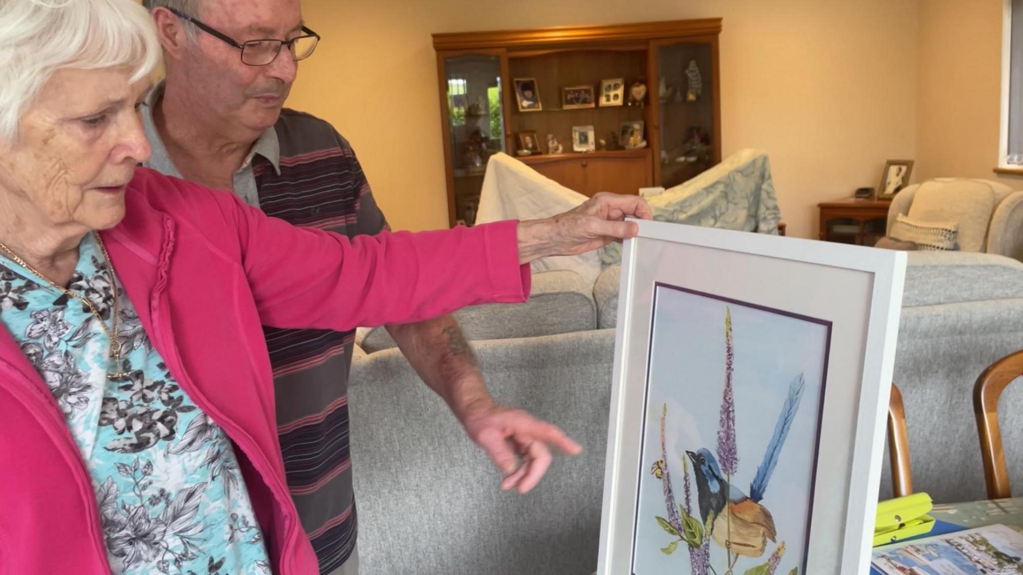 Jill Bristow and her husband George look at a painting of a bird perched on some flowers in their home. Jill is wearing a pink fleece and a blue T-shirt with grey flowers on it. George has a brown, grey and pink striped T-shirt on.