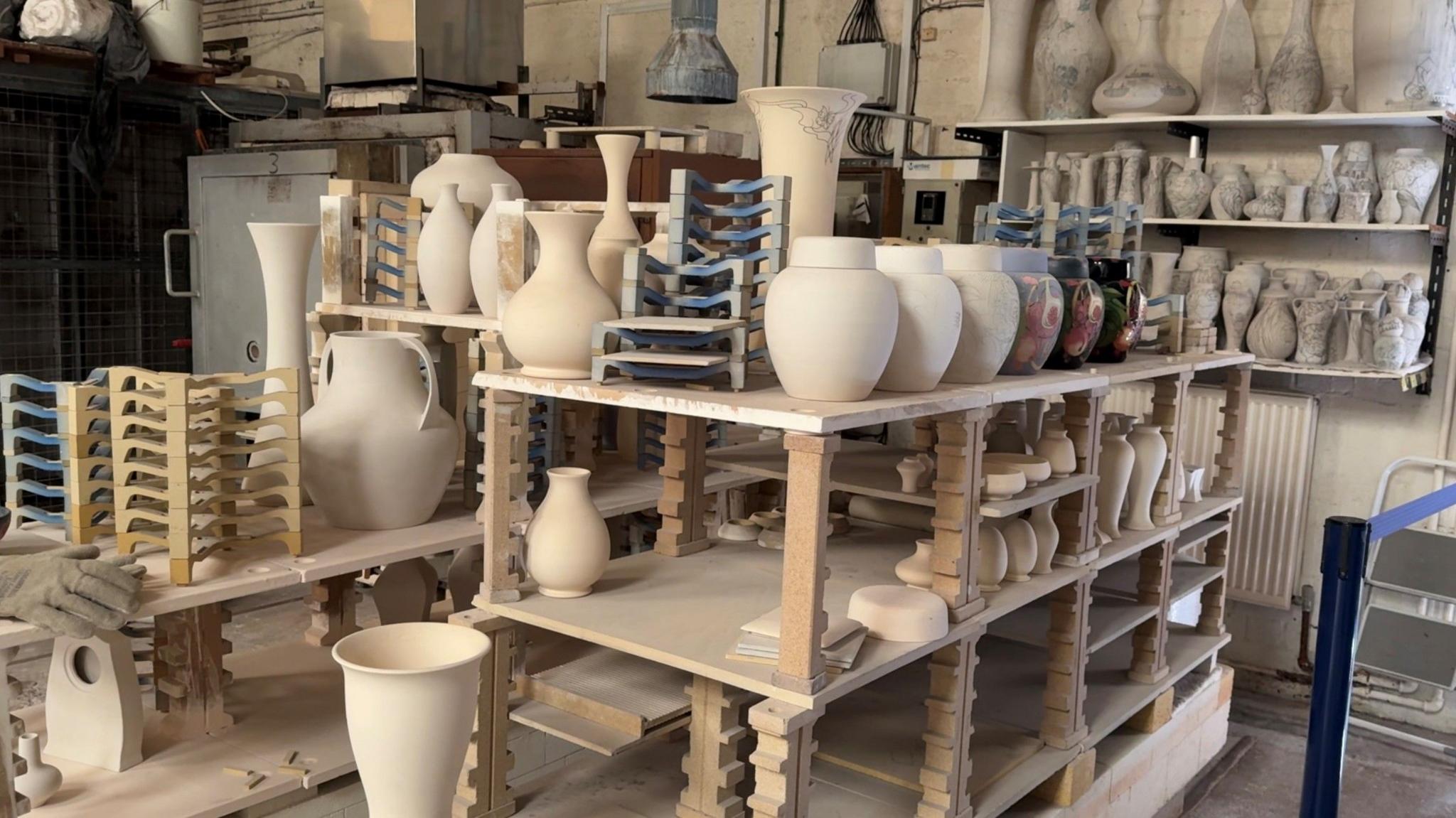 Shelving which contains unpainted Moorcroft pottery in a warehouse. There are various different shapes and sizes. There is a set of products in a line which illustrates the manufacturing process.