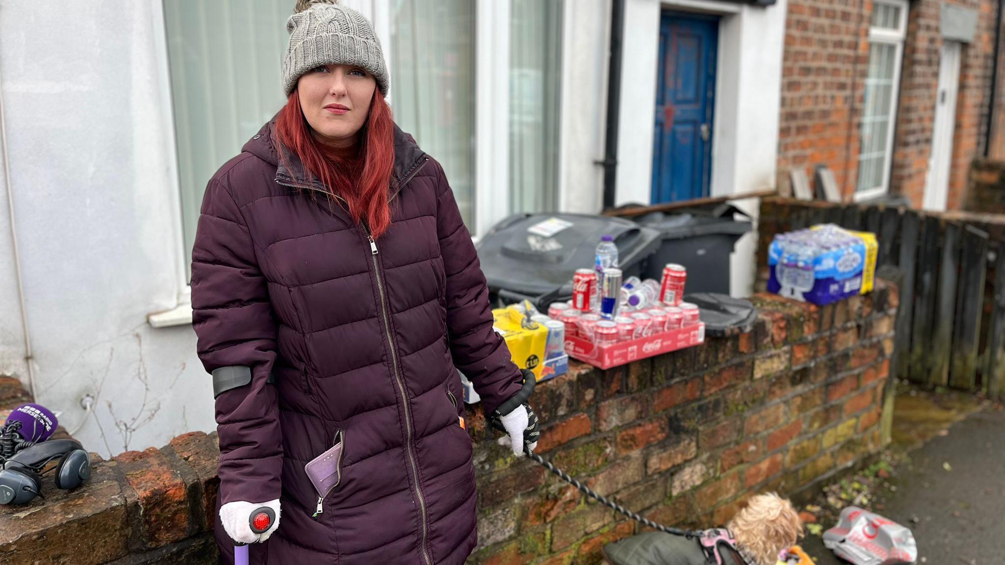 Laura Caunce, who has long red hair and is wearing a purple coat and a grey woolly hat, poses for the camera holding her Yorkshire Terrier's lead in her left hand. There are cans of Coca-Cola and Red Bull and bottles of water on a garden wall behind her