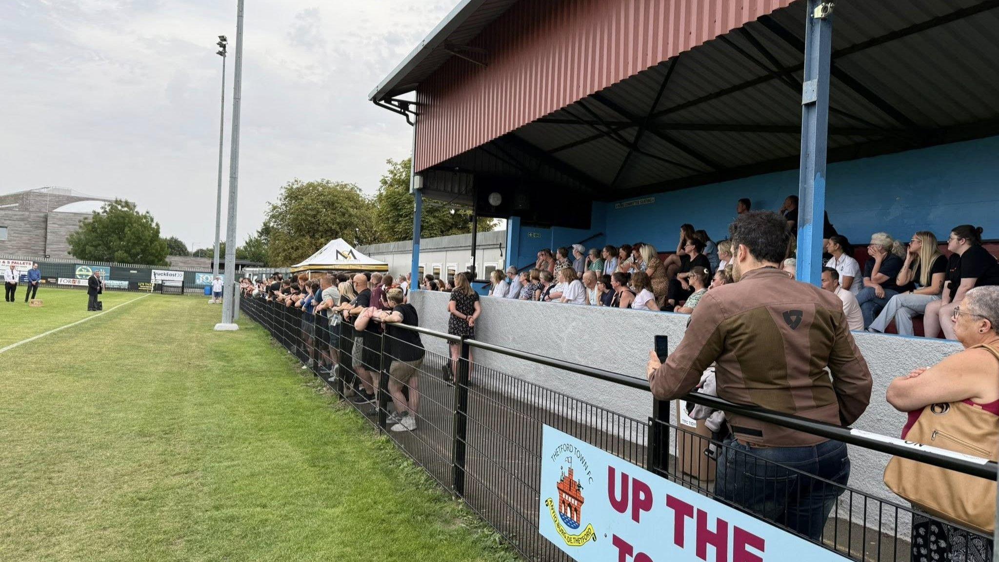 A small group of people stood on a football pitch address a crowd of about 300 people gathered behind fencing and in a stadium stand at an event.