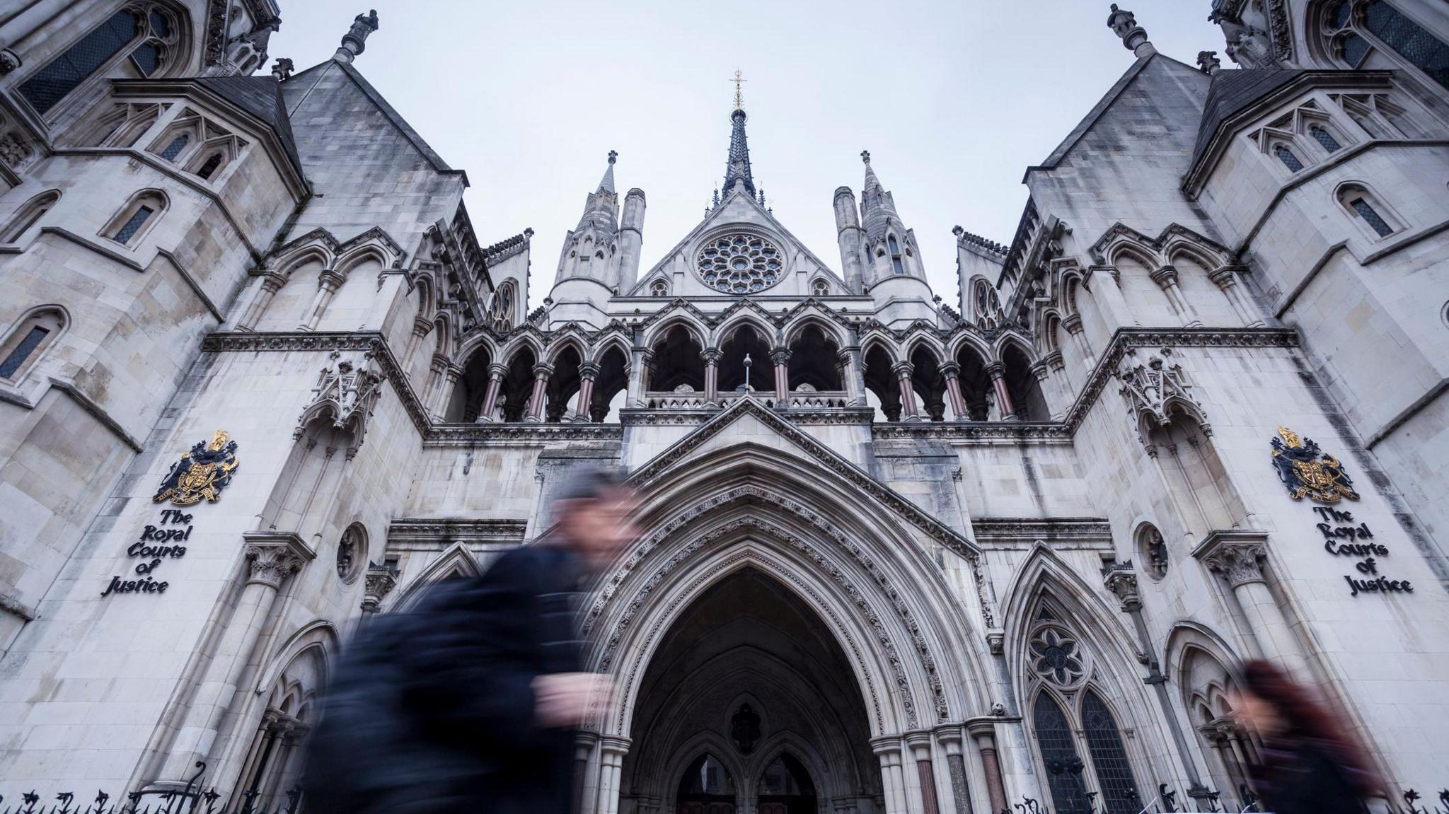 An exterior view of the Royal Courts of Justice, taken from a low viewpoint, showing the neo-Gothic arches and spires reaching towards the sky as figures blurred by their movement pass in front of the building