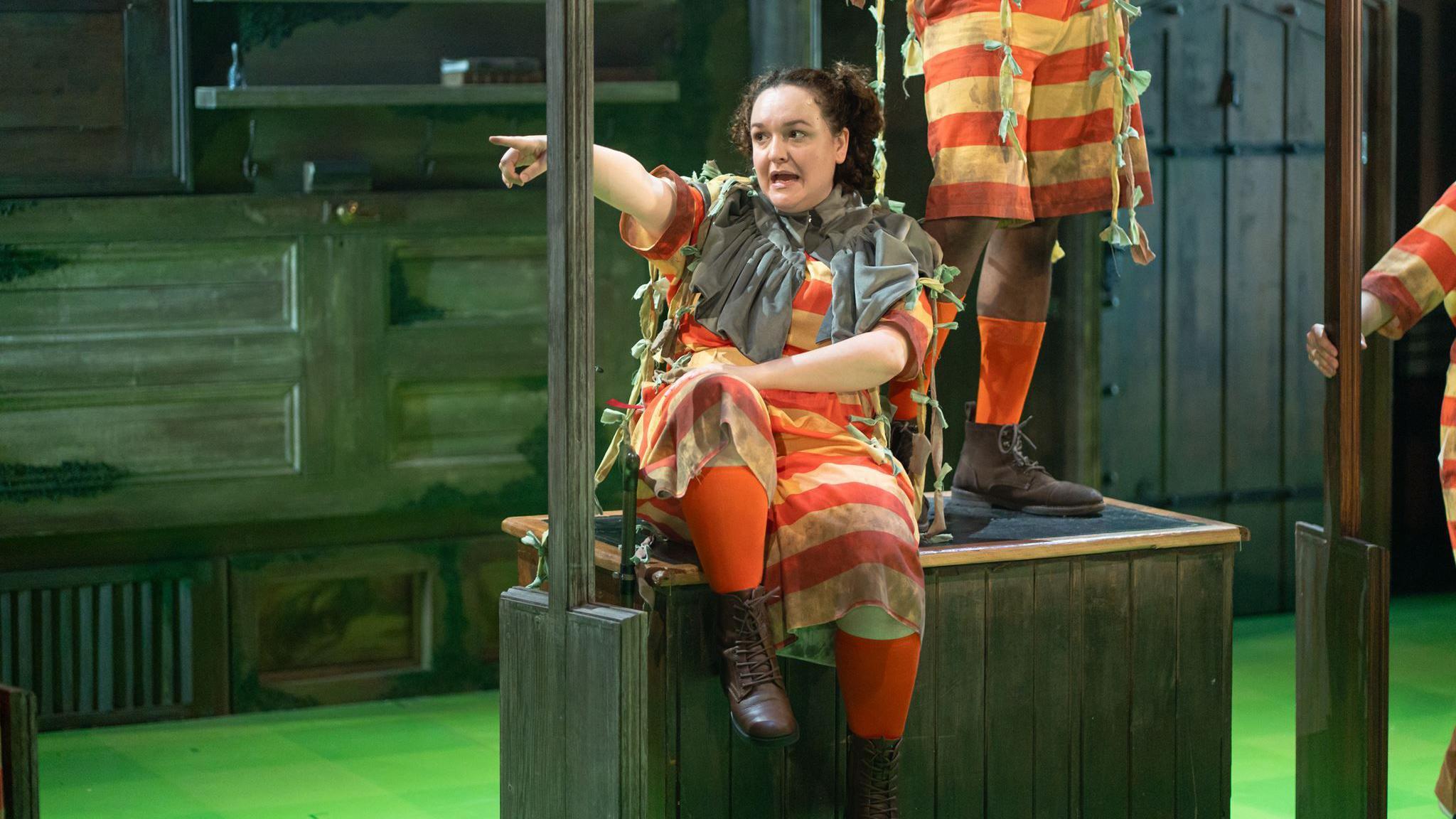Daneka Etchells performs in a red-and-orange striped costume. She is sitting on a wooden platform and pointsingacross a theatre stage. Other cast members in matching costumes are partially visible among dark wooden set pieces under green stage lighting.