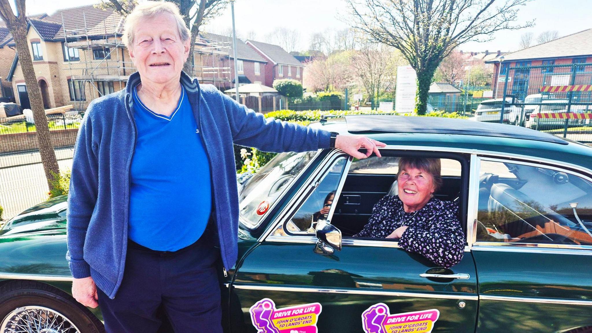 Dave Whittington wearing a blue t-shirt and cardigan, standing near the passenger door of his dark green 57-year-old MGC classic car, with his wife Thelma Whittington sitting in the car.