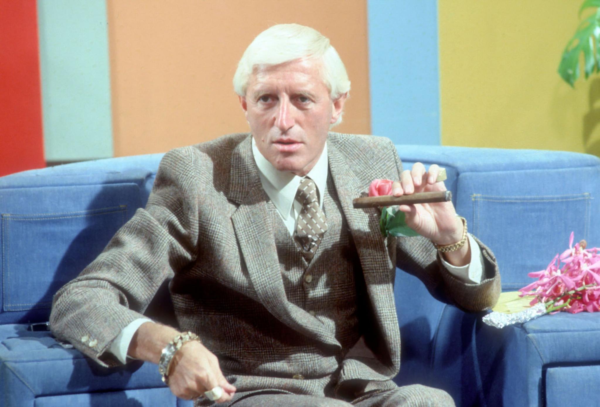 Jimmy Savile smoking a cigar and wearing a grey suit, sitting on a blue sofa