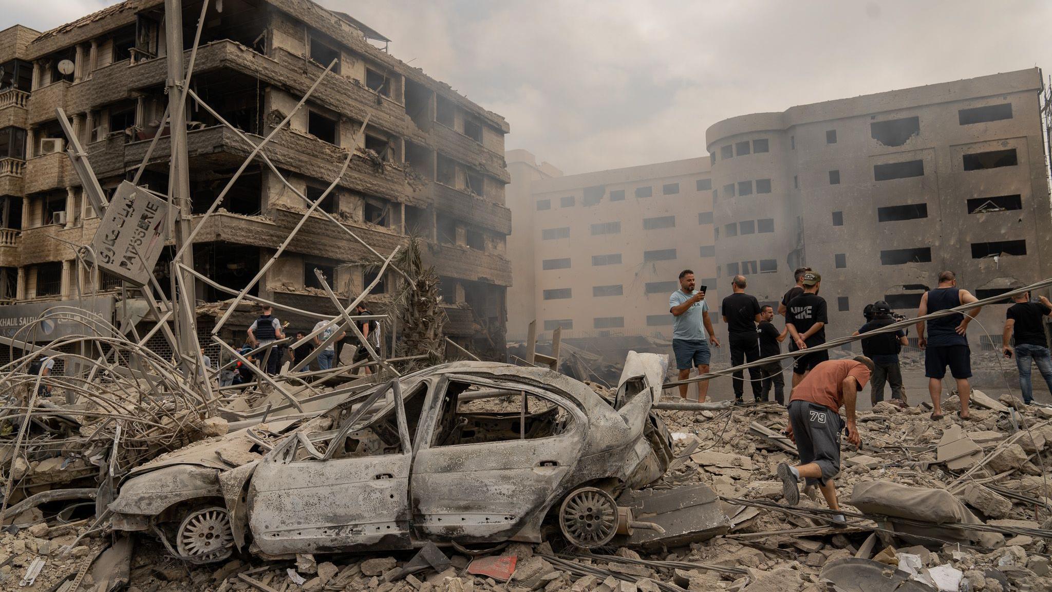 A car is coated completely in grey dust and surrounded by rubble, while a group of men survey the scene over damaged buildings.