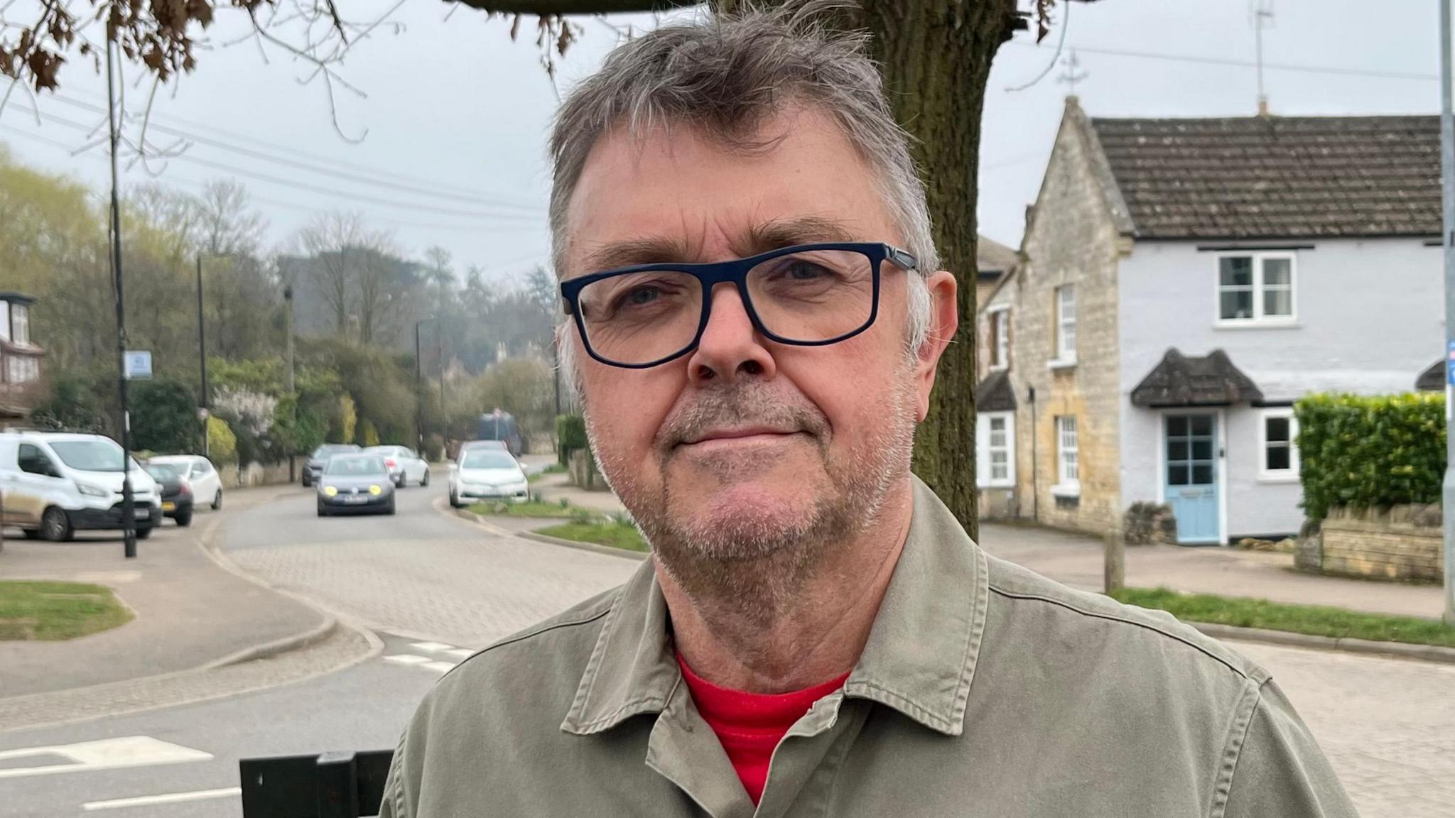 Martin Chillcott, 68, has brown hair with greying hair on each side. He is wearing a khaki shirt with an orange T-shirt under that. He has a large badge on the left of his shirt that says Protect Rural Peterborough. Roadways are visible behind him, with cars parked along near the kerbs. Some daffodils are in bloom on the left and a tree is behind him. There are some houses each side of the road.