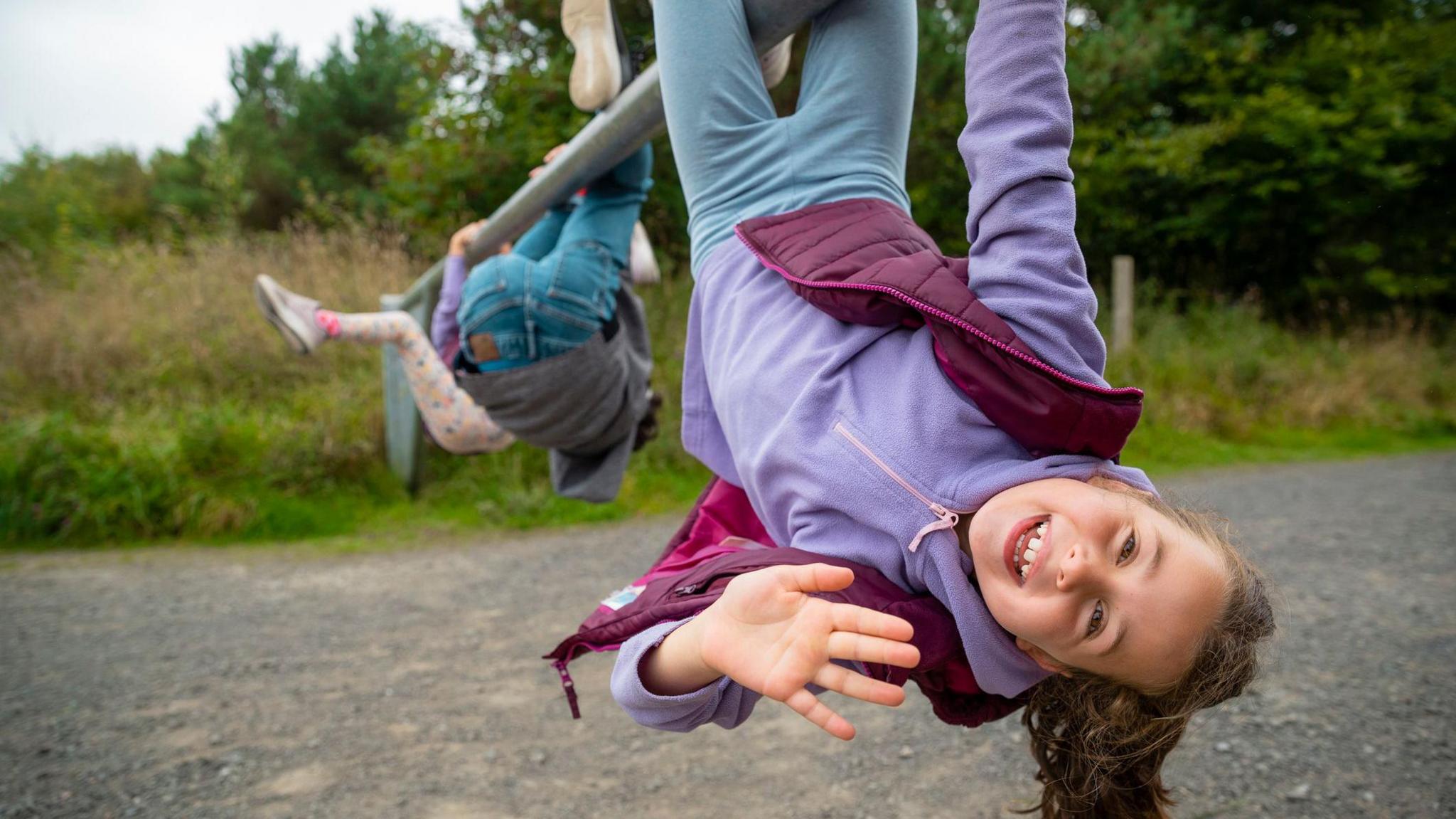 A girl hangs upside down from a metal bar. She is smiling and waving at the camera. There are two children in the background doing the same but facing away from the camera