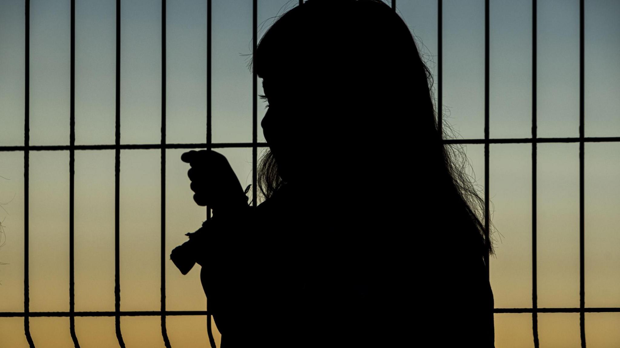 Young girl looks through metal fence, at sunset. She is silhouetted.