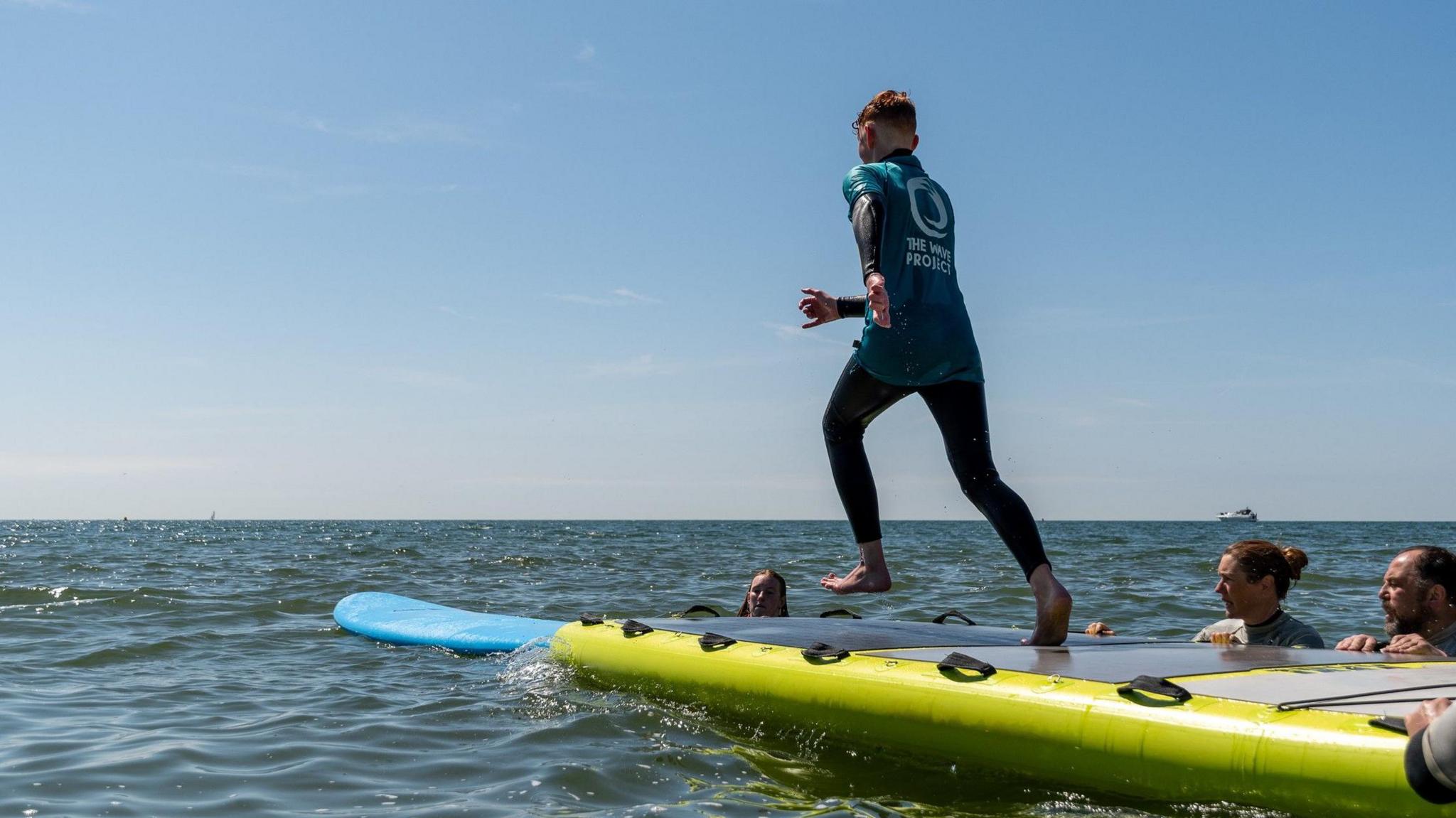 A boy jumping off a mini-jetty into the sea with volunteers around and supporting him 